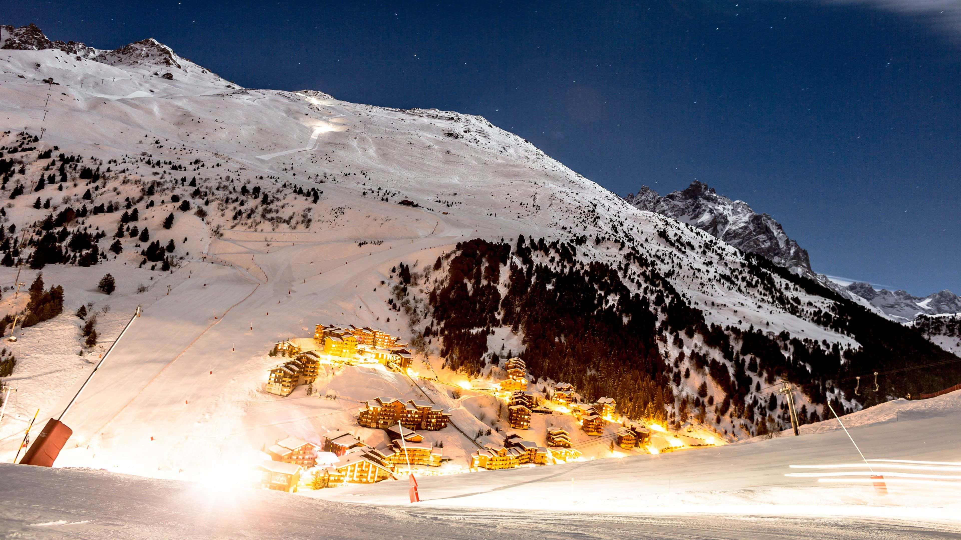 Ski Resort 桌布 壁紙 — 一個被雪覆蓋的山村在星空下溫暖地發光，滑雪纜車和小徑蜿蜒穿過山峰。