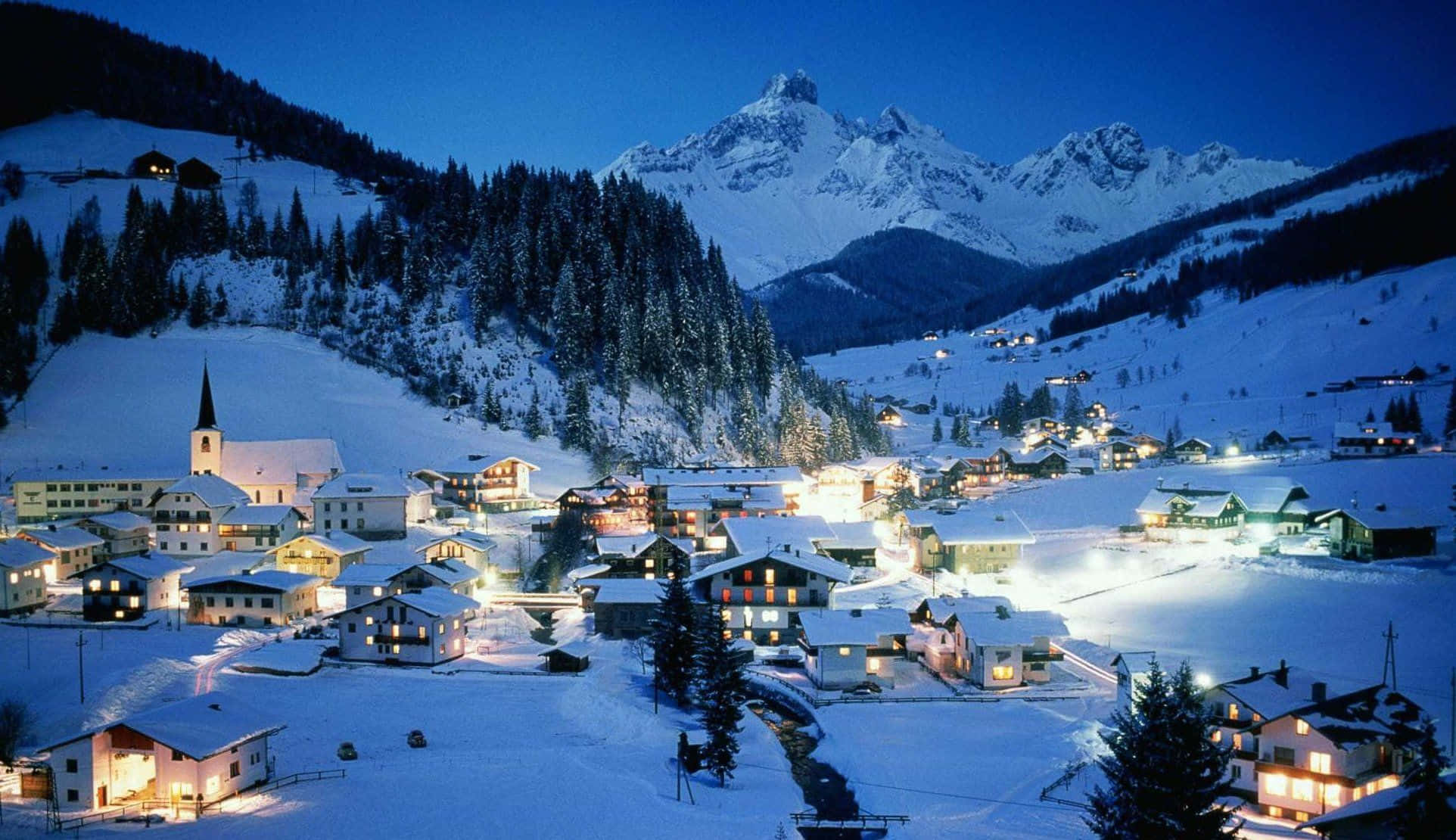 Ski Resort 桌布 壁紙 — 一個被雪覆蓋的山村在黃昏時分溫暖地發光，窗戶和教堂尖塔的燈光映襯著深藍色的天空。