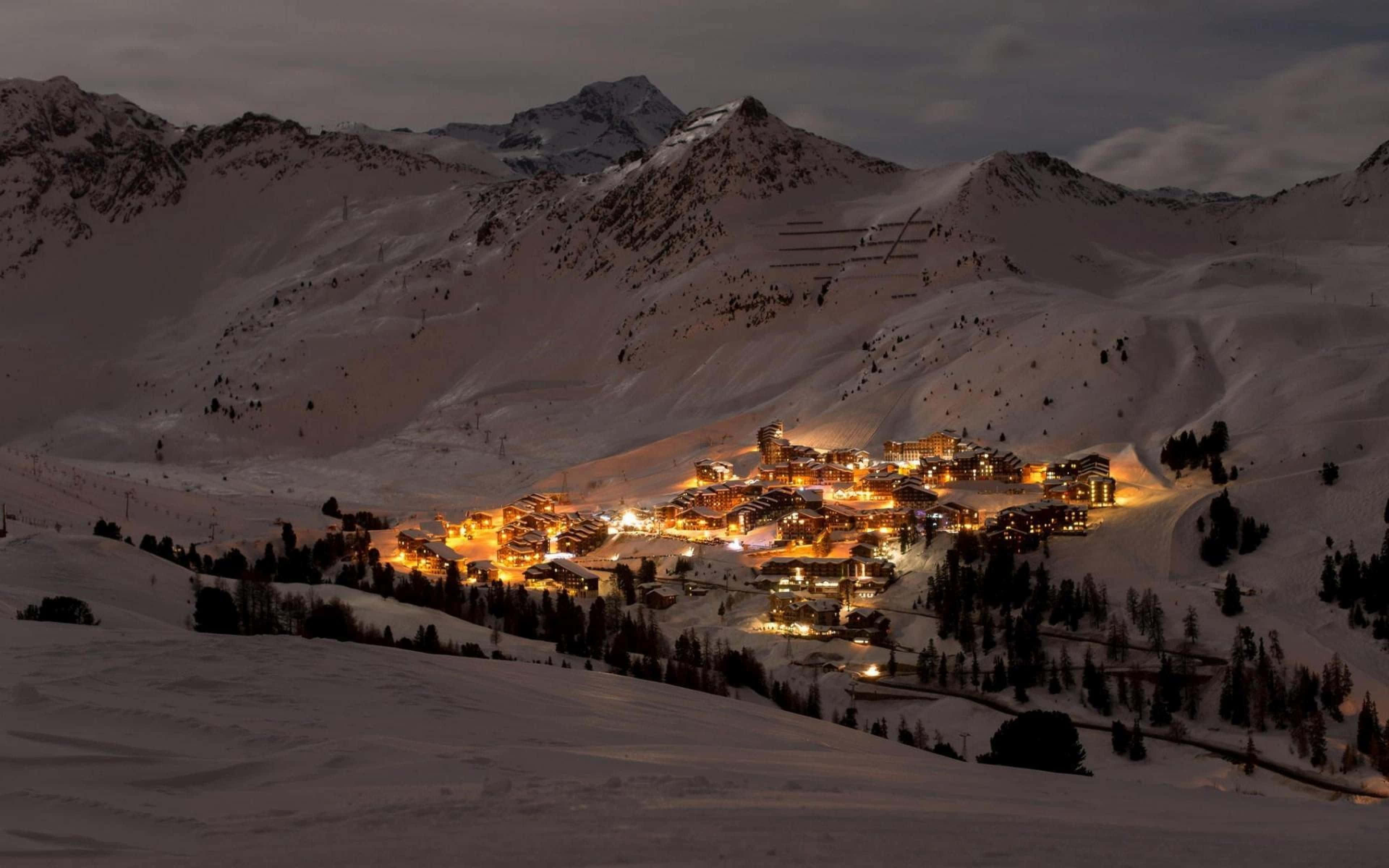 Snowy Village 桌布 壁紙 — 一個被雪覆蓋的山村在黃昏時分溫暖地發光，坐落在多雲天空下高聳的山峰之間。