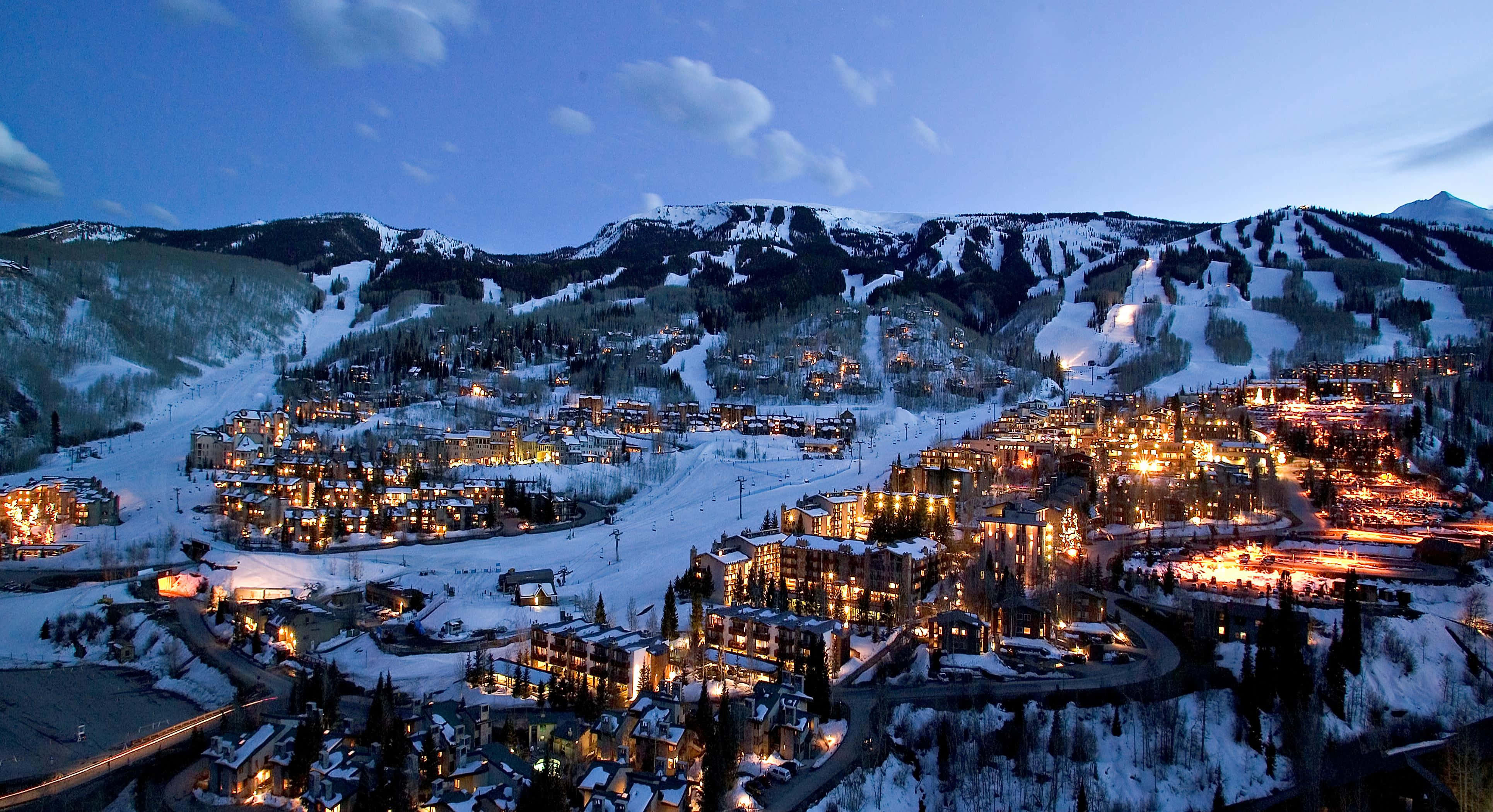 Aspen Colorado 桌布 壁紙 — 一個被雪覆蓋的山村在黃昏時溫暖地發光，燈火通明的建築物坐落在滑雪道之間，在暮色天空下。