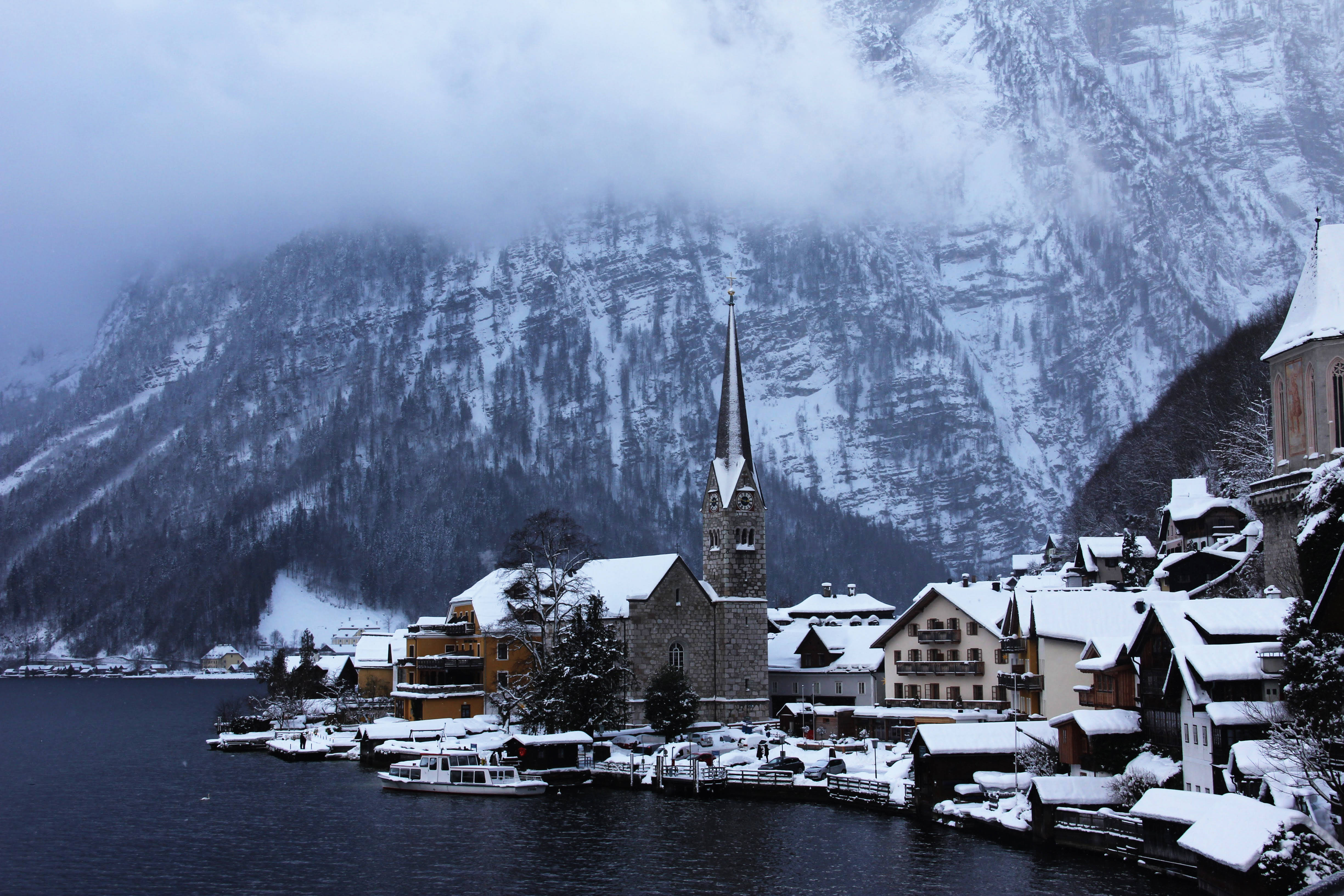 Austria 桌布 壁紙 — 一個被雪覆蓋的村莊緊貼湖岸，石砌教堂的尖頂在霧氣繚繞、森林覆蓋的山巒中聳立。