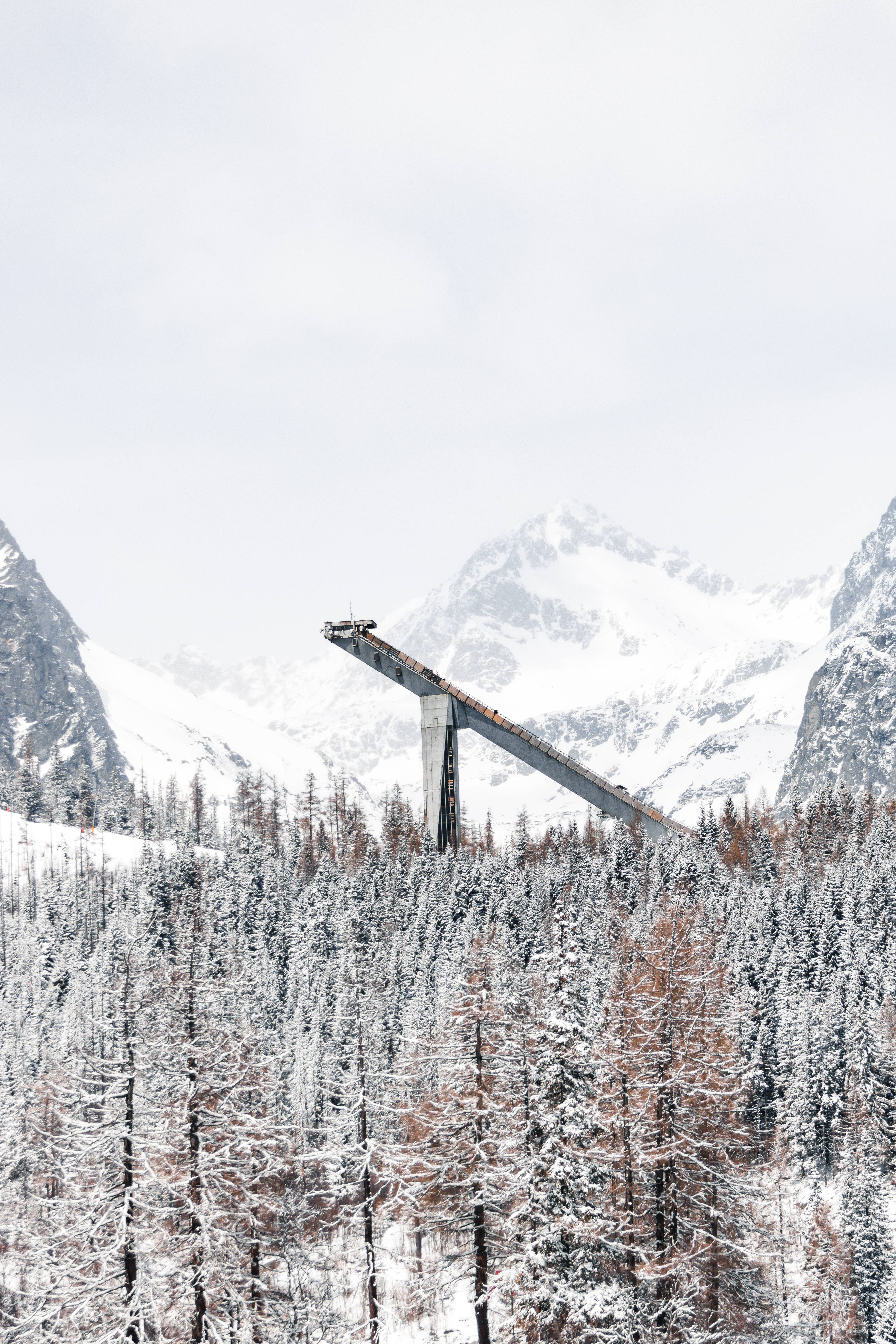 Slovakia 桌布 壁紙 — 一個被雪覆蓋的滑雪跳台從森林覆蓋的山谷中戲劇性地升起，背景是陰天下一座座高聳的雪山。