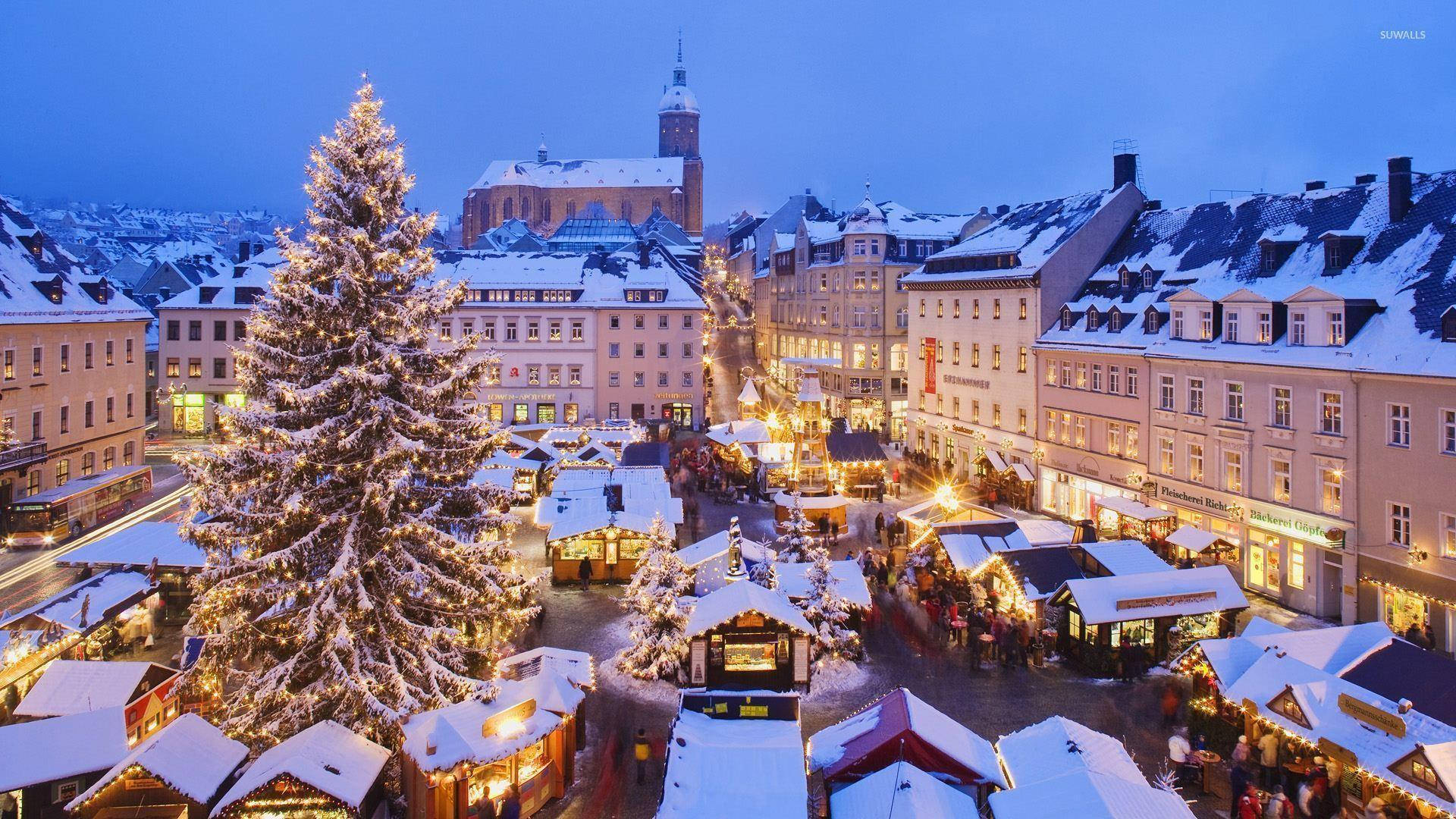 Munich 桌布 壁紙 — 一個被雪覆蓋的聖誕市集在暮色中溫暖地閃耀，背景是高聳的樹木和歷史建築。