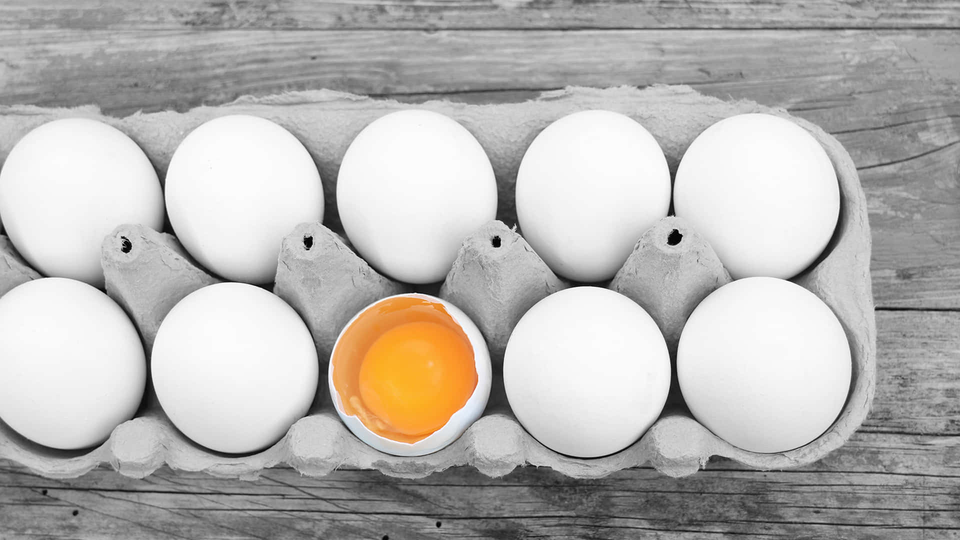 Eggs 桌布 壁紙 — 一個裂開的雞蛋帶有鮮橙色蛋黃，在灰色調的雞蛋盒中脫穎而出，置於風化木頭上。