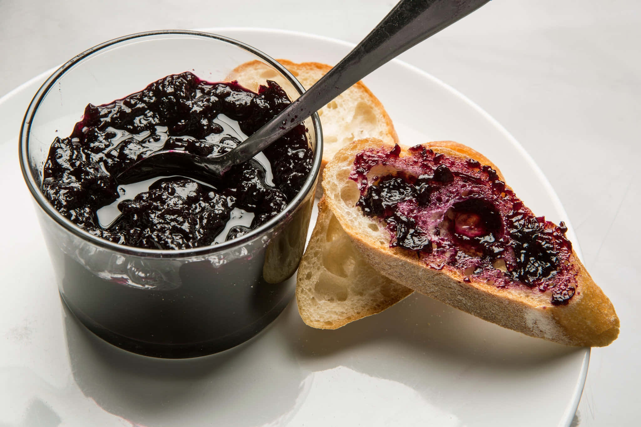 Blueberry Jam 桌布 壁紙 — 一個裝有深紫色莓果醬的玻璃罐和湯匙放在白色盤子上，旁邊是一片塗了果醬的脆皮麵包。