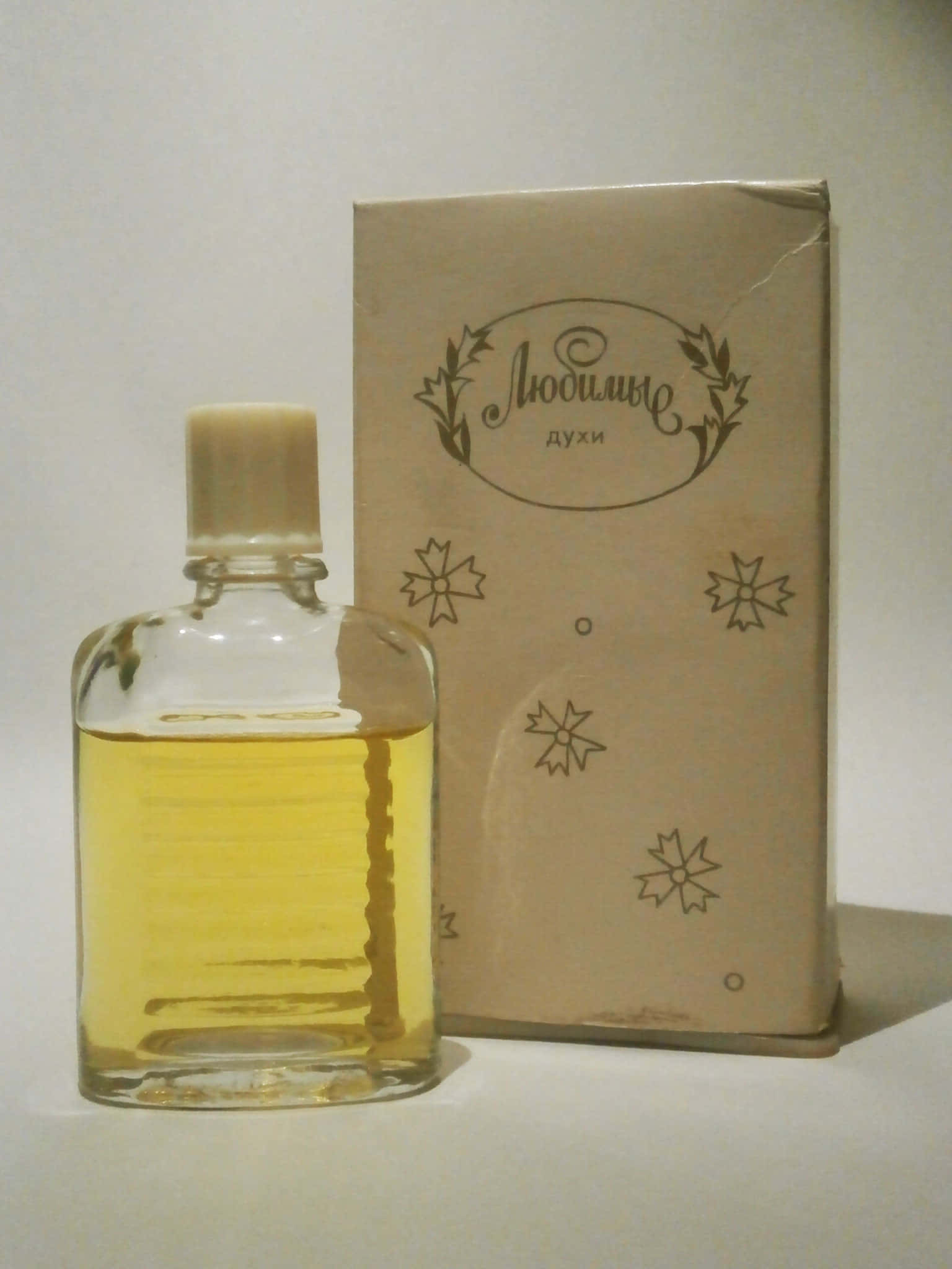 perfume 桌布 壁紙 — 一個裝有琥珀色液體的復古玻璃香水瓶，旁邊放著它的米色盒子，盒子上飾有花卉字體和星爆圖案。