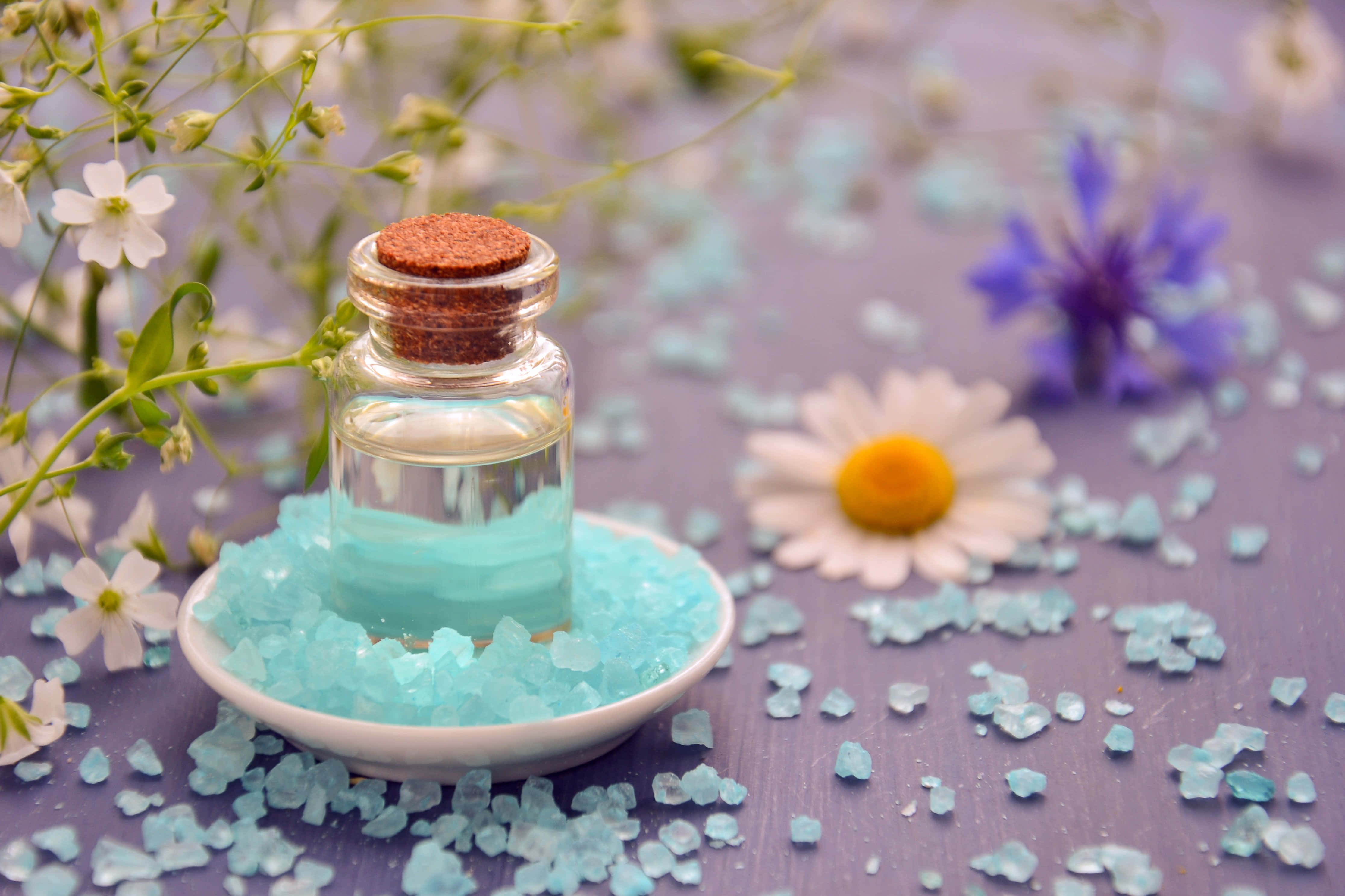 Aromatherapy 桌布 壁紙 — 一個裝有藍色液體的玻璃瓶，軟木塞蓋著，放在白色碟子中，周圍環繞著藍色玻璃珠和精緻的白色和紫色花朵，置於紫色表面上。