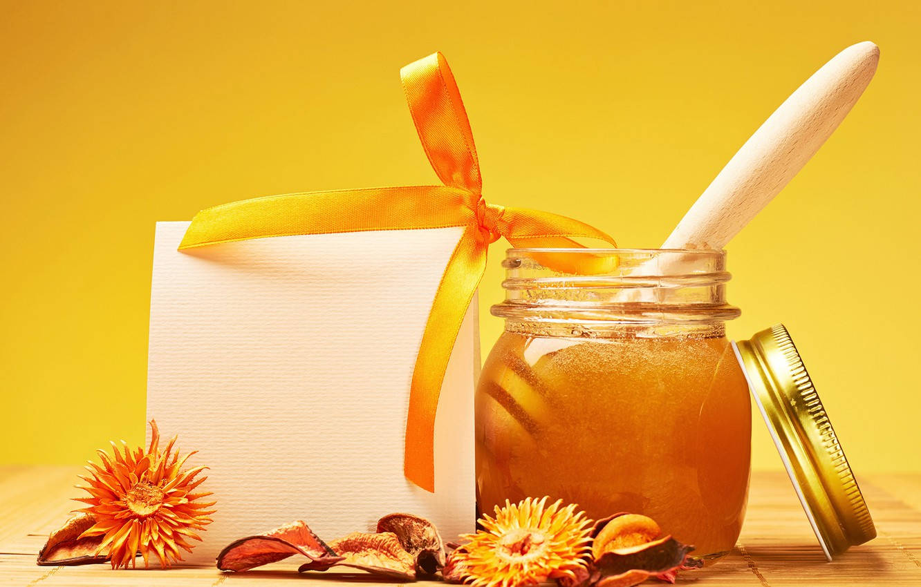 honey 桌布 壁紙 — 一個裝有金色蜂蜜的玻璃罐和一個木勺，旁邊放著一個用黃色絲帶和乾花裝飾的禮物盒，放在木質表面上。