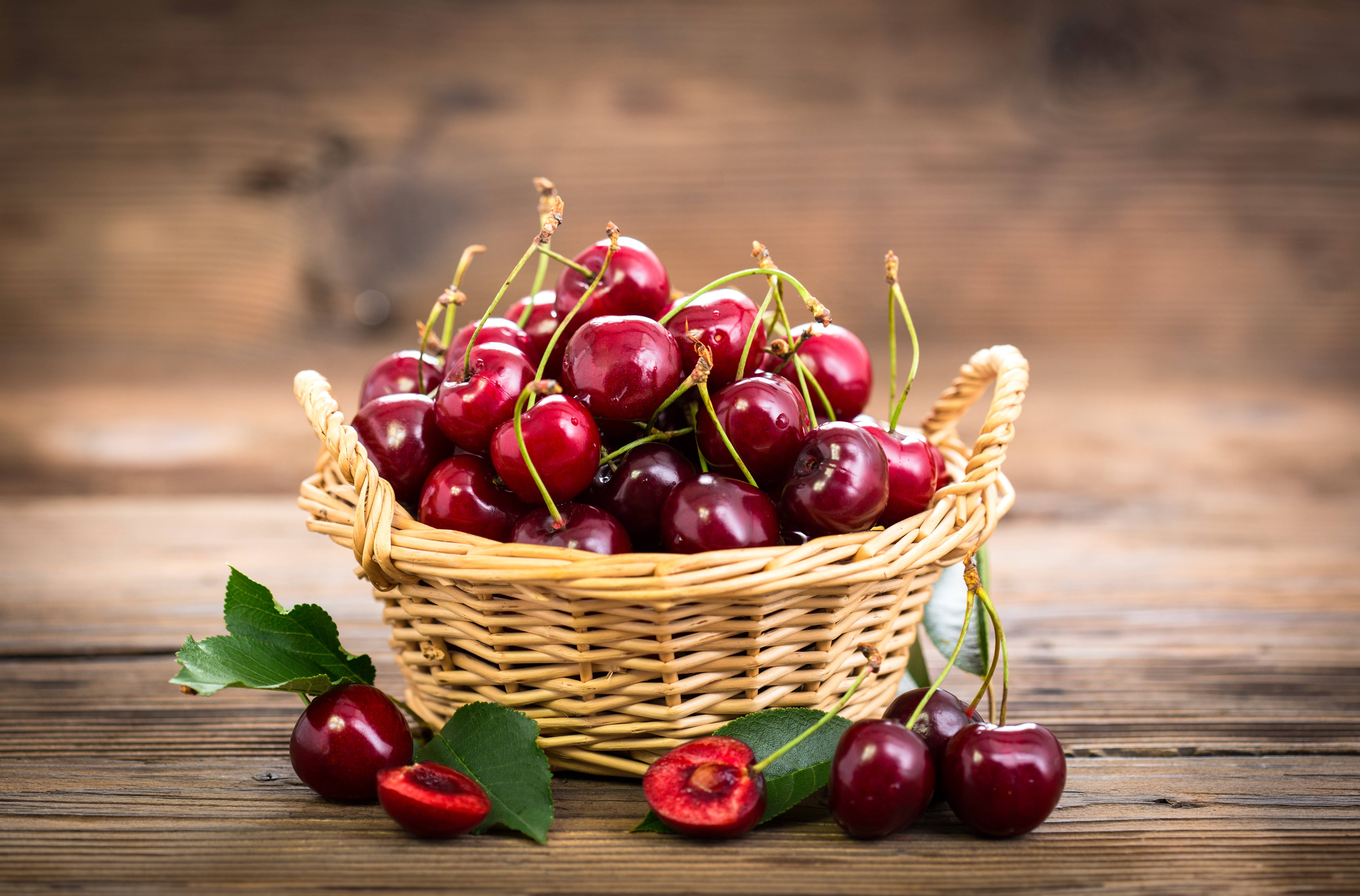 Cherry 桌布 壁紙 — 一個裝滿光澤紅櫻桃的籃子放在質樸的木桌上，周圍散落著樹葉和半個櫻桃。