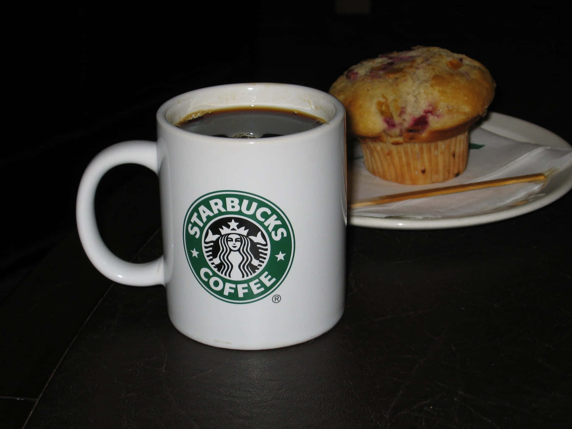星巴克 桌布 壁紙 — 一個裝滿咖啡的白色Starbucks馬克杯放在深色桌子上，旁邊有一個莓果鬆餅，光線昏暗。
