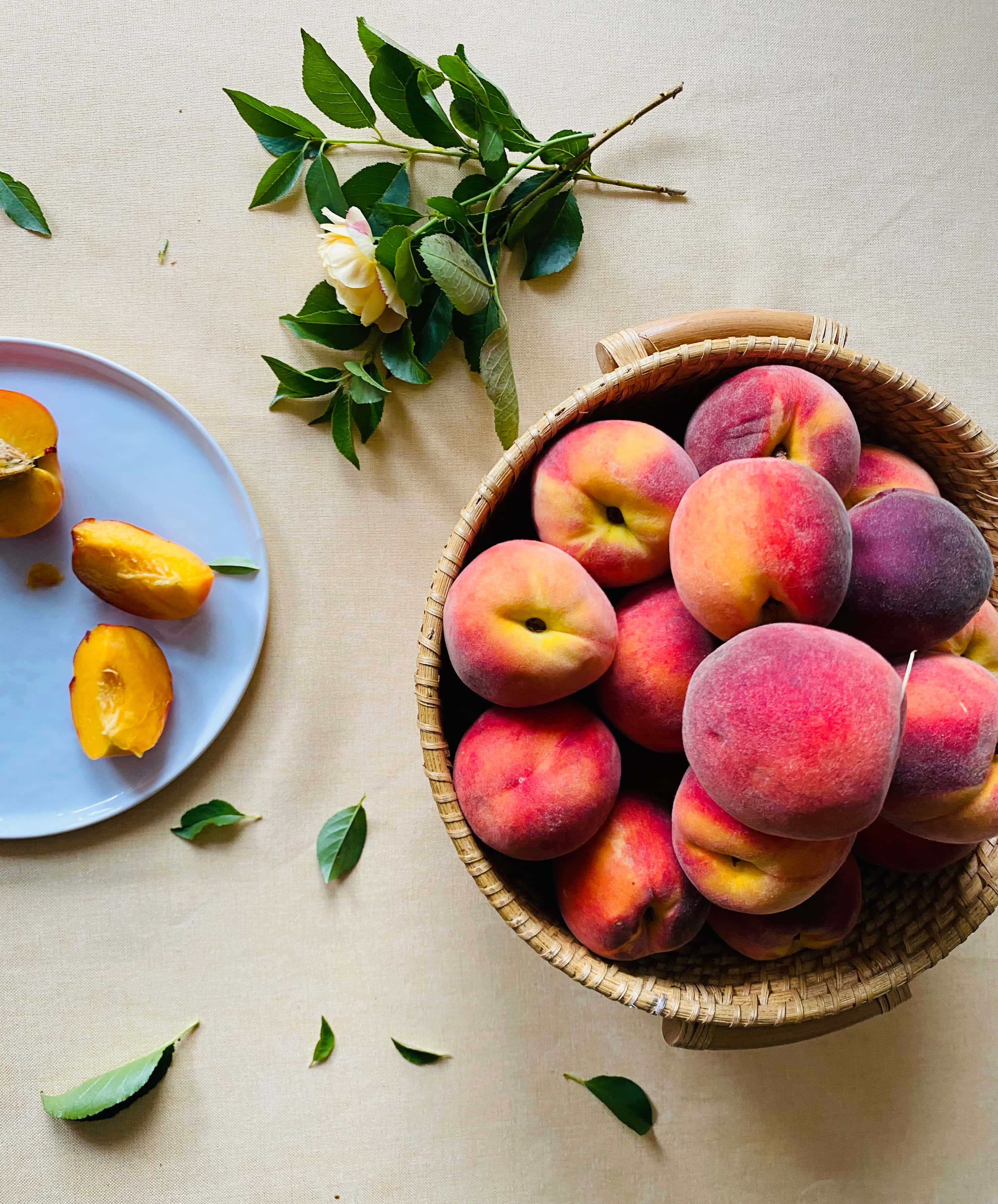 Peaches 桌布 壁紙 — 一個裝滿成熟桃子的編織籃子旁邊放著一個盤子，裡面有切半的水果和一朵玫瑰枝，放在米色亞麻布面上。
