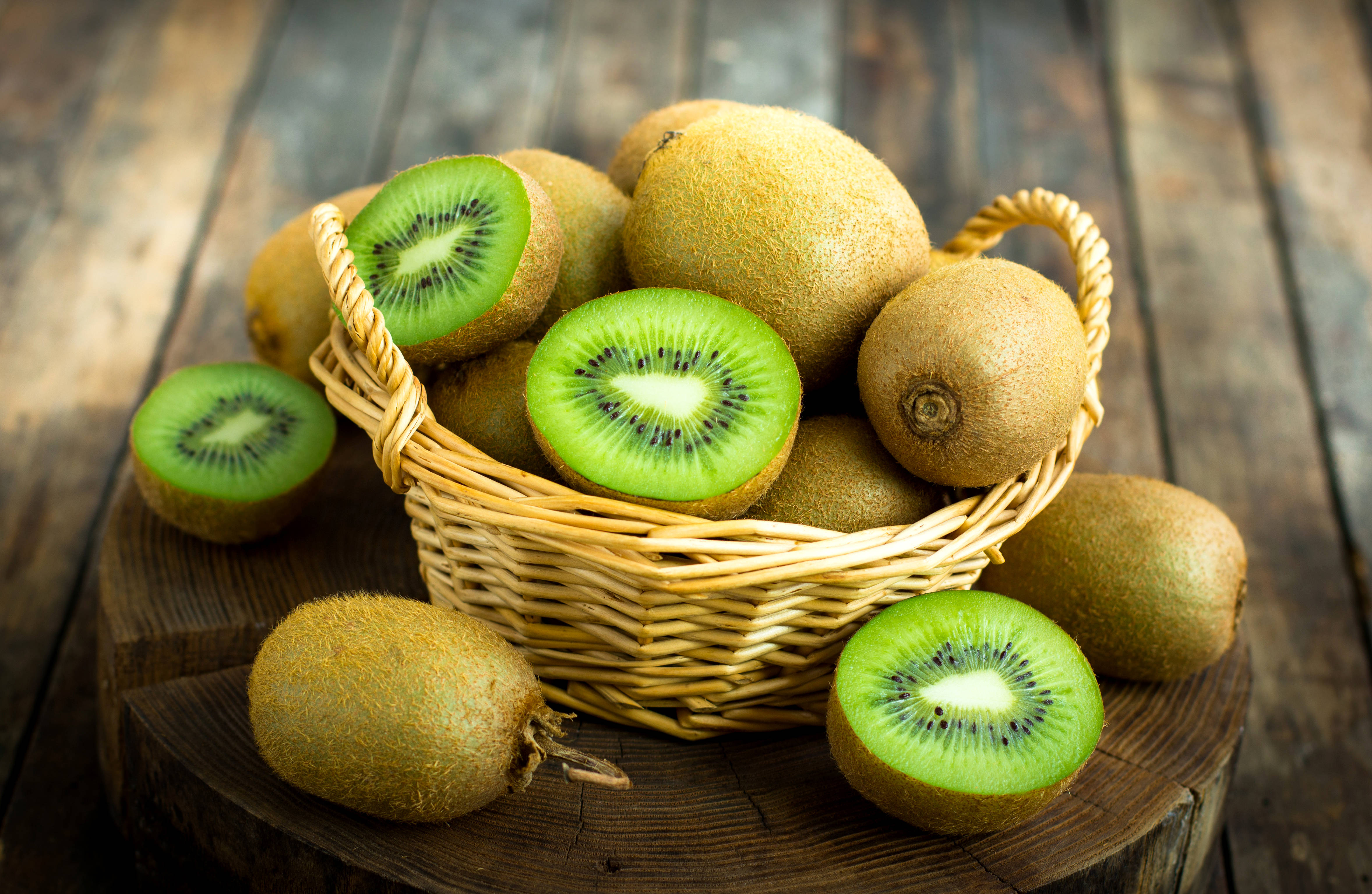 Kiwi 桌布 壁紙 — 一個裝滿新鮮奇異果的籃子，有些切片露出鮮綠色果肉和黑色種子，放在質樸的木桌上。