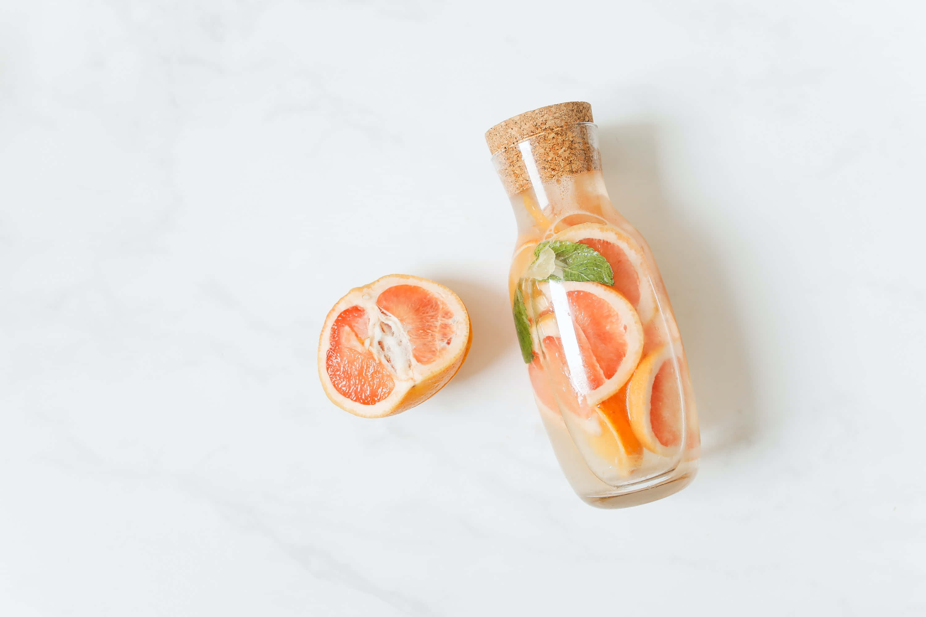 Summer Drinks 桌布 壁紙 — 一個裝滿柑橘片和薄荷的玻璃瓶放在白色大理石表面上，旁邊是一個切半的葡萄柚。