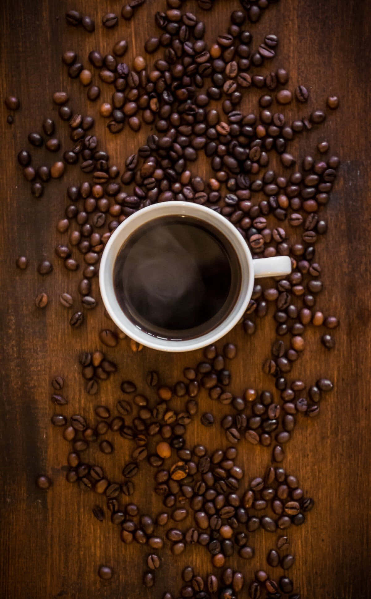 Coffee Cup 桌布 壁紙 — 一個裝滿深色咖啡的白色咖啡杯，周圍散落著烘焙咖啡豆，放在質樸的木質表面上。