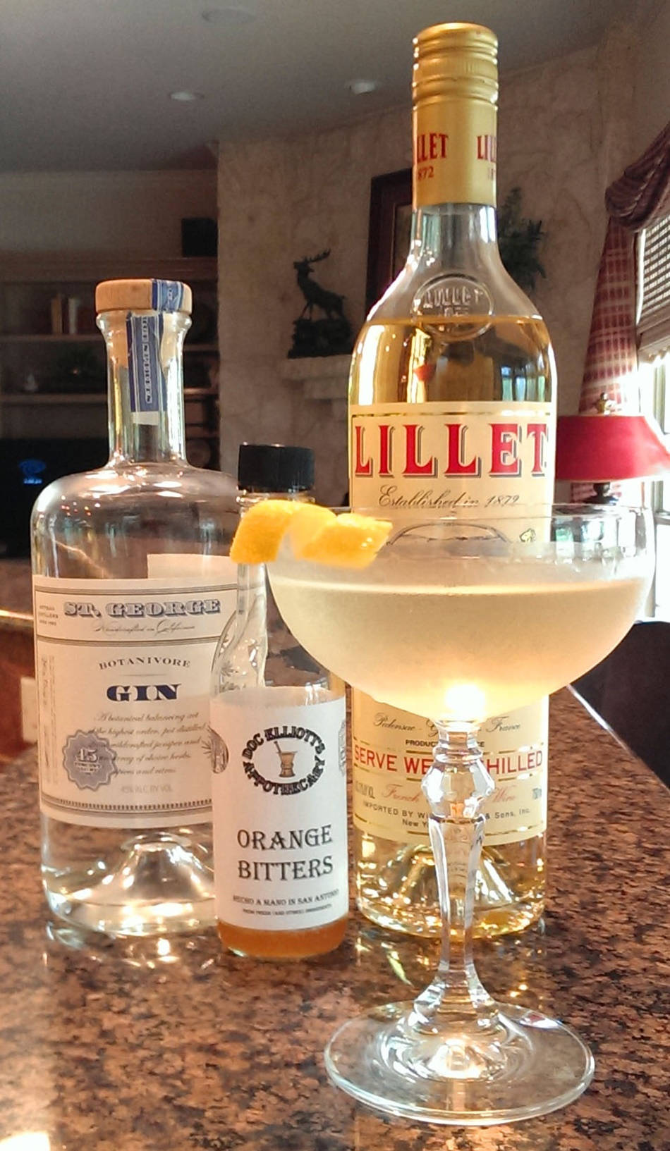 drinks 桌布 壁紙 — 一個裝滿淺色液體和橙裝飾的雞尾酒杯，旁邊放著琴酒苦精和利萊酒的瓶子，在一個有斑點的櫃檯上。