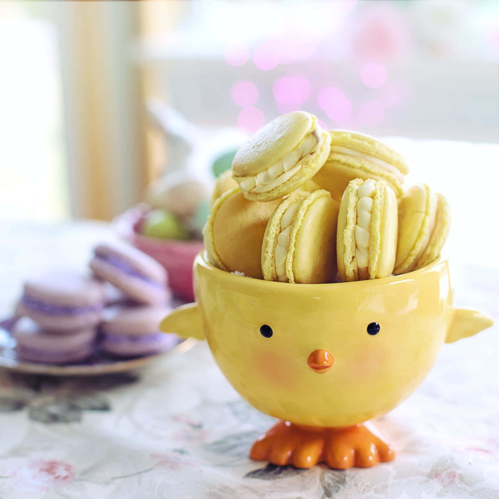 Pastel Easter 桌布 壁紙 — 一個裝滿淺黃色馬卡龍的開心黃色小雞形碗，放在花卉桌布上，背景有模糊的粉彩馬卡龍。