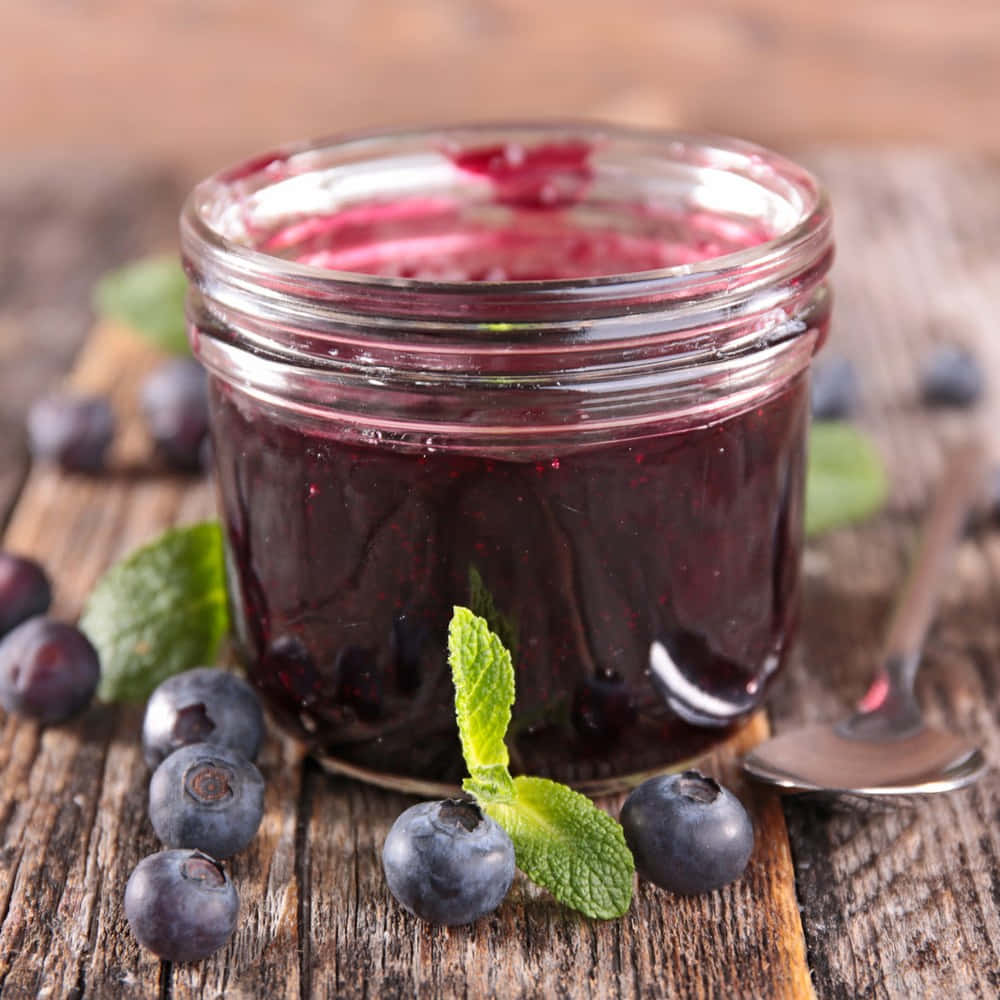 Blueberry Jam 桌布 壁紙 — 一個裝滿濃郁深藍色藍莓醬的玻璃罐放在質樸的木桌上，周圍是新鮮藍莓和薄荷葉。
