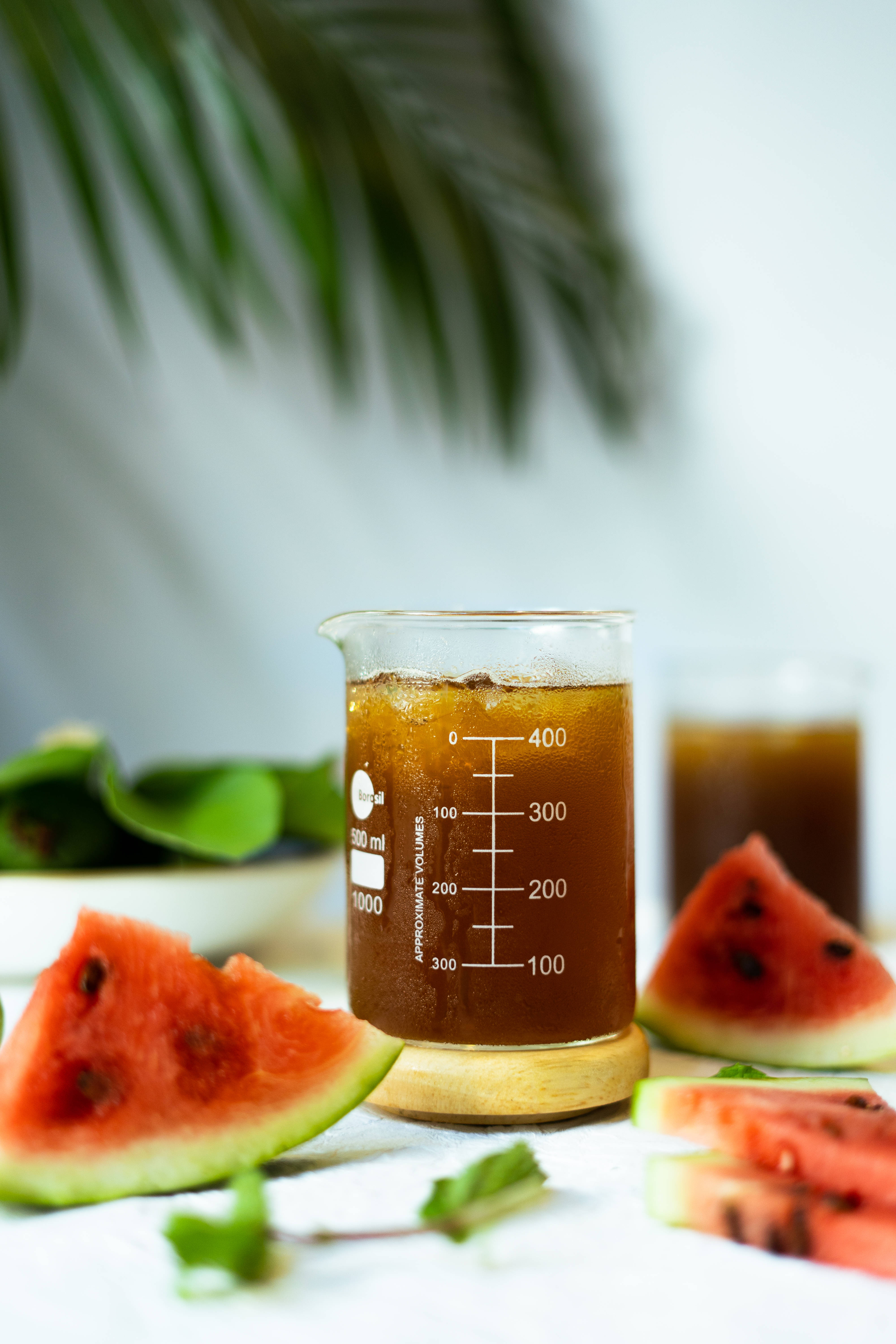 Watermelon 桌布 壁紙 — 一個裝滿琥珀色液體的玻璃燒杯，周圍環繞著鮮豔的西瓜片和綠葉，背景是柔和的白色。