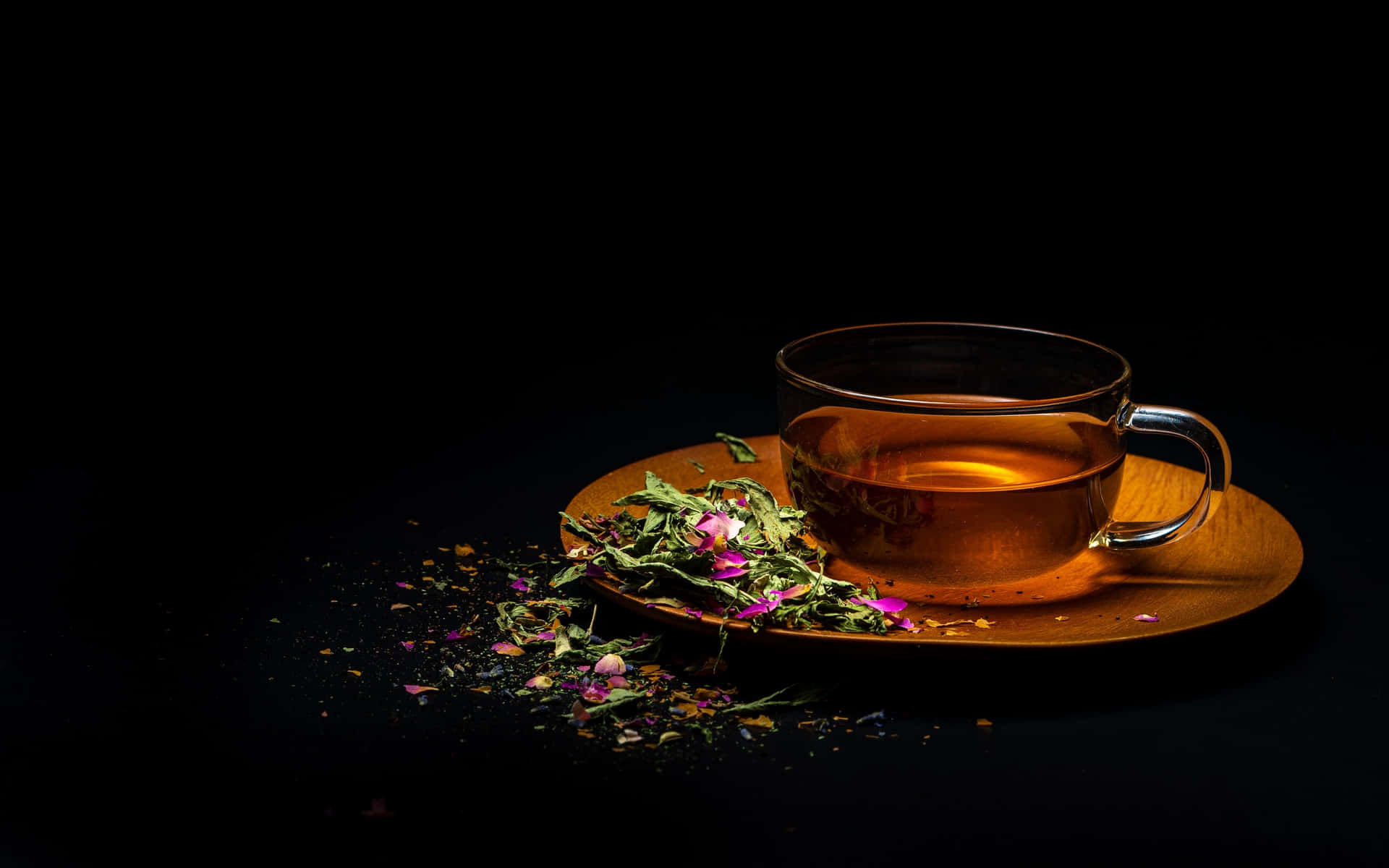 咖啡動圖 桌布 壁紙 — 一個裝滿琥珀色茶的玻璃茶杯放在木製茶托上，周圍散落著乾燥的玫瑰花瓣和葉子，背景為黑色。