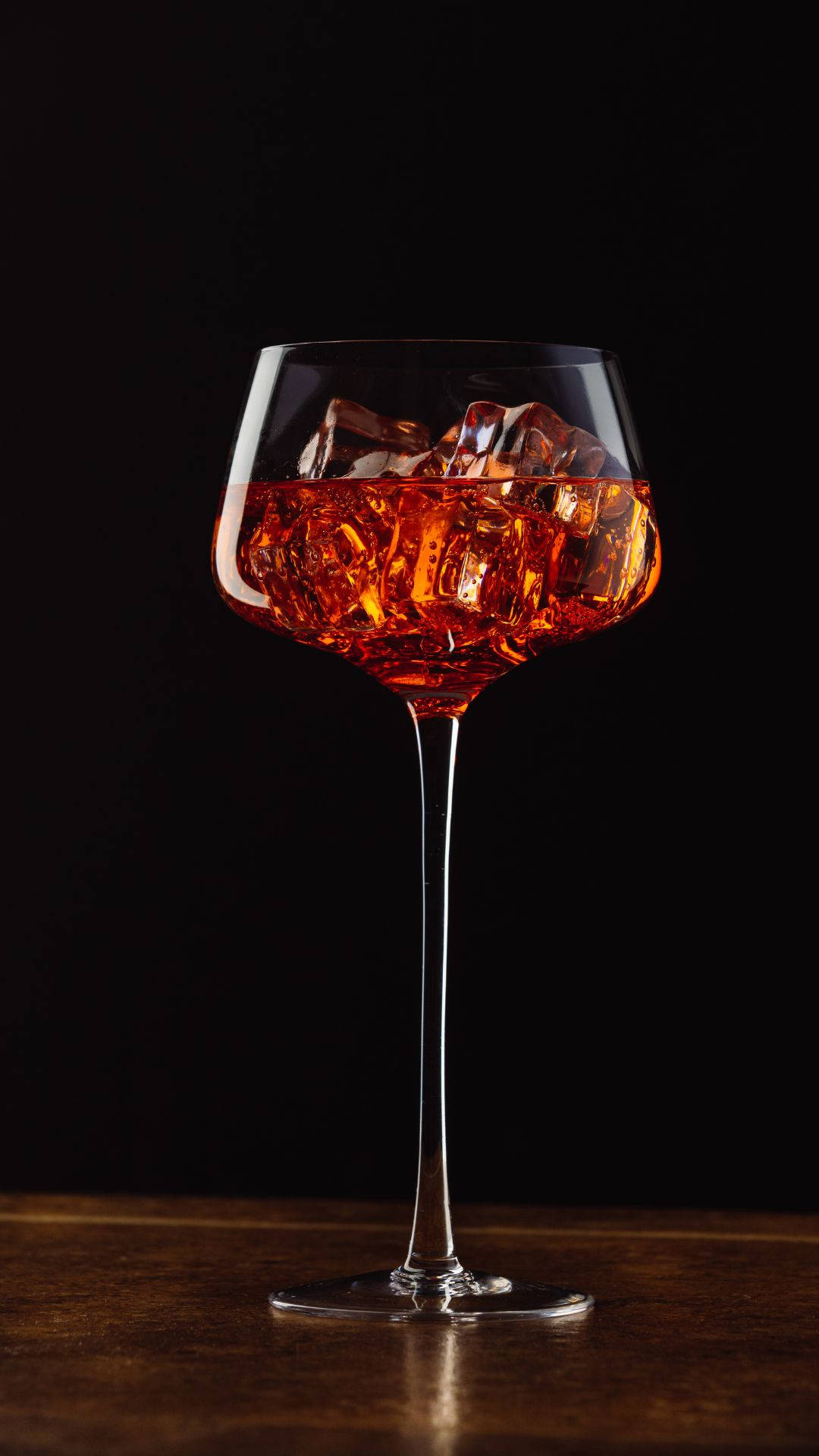 alcohol 桌布 壁紙 — 一個裝滿琥珀色雞尾酒和冰塊的有腳玻璃杯在黑色背景下的木質表面上閃耀。
