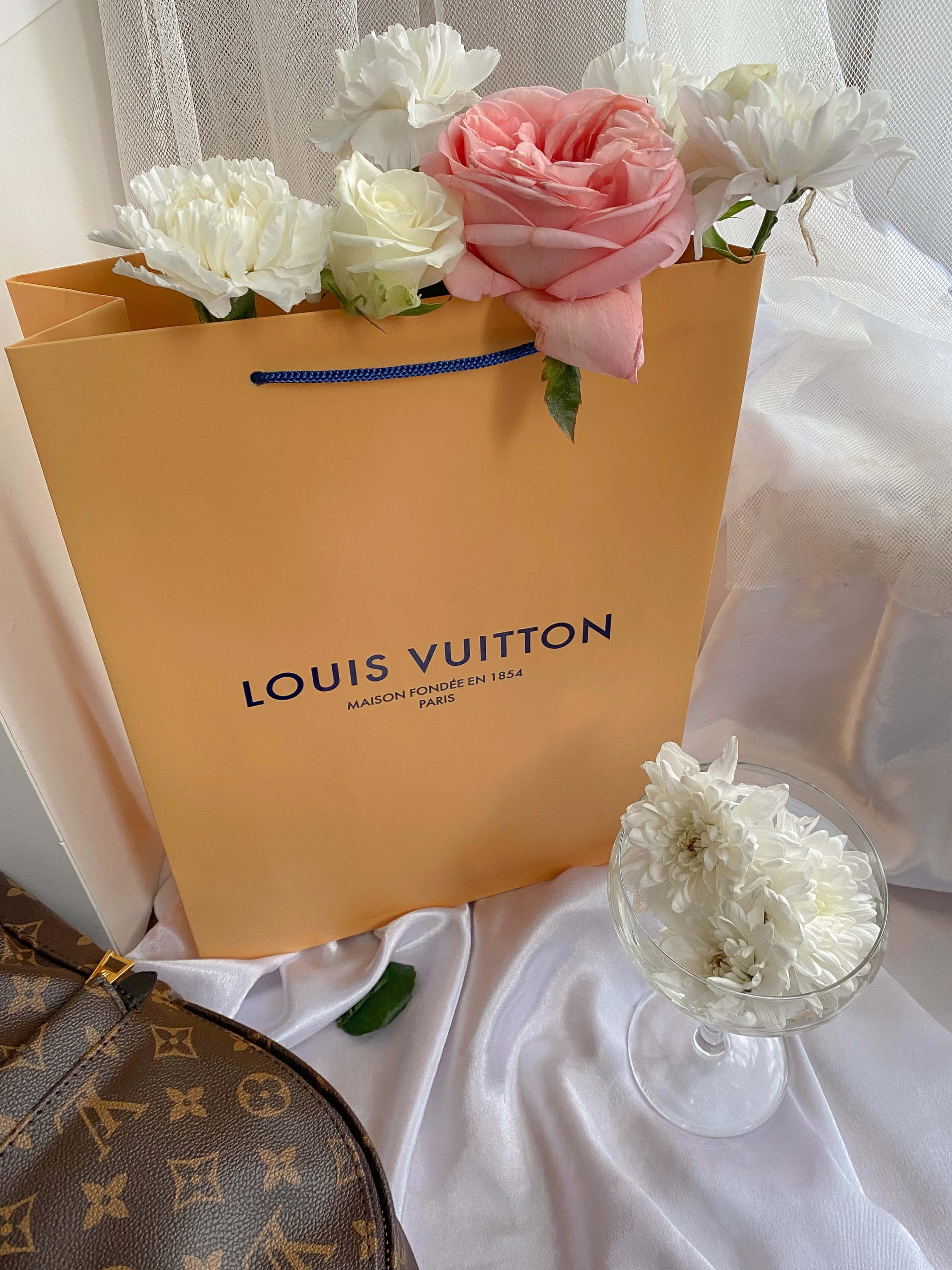 路易威登 桌布 壁紙 — 一個裝滿粉紅色和白色花朵的Louis Vuitton購物袋放在白色緞面表面上，旁邊是一個裝有白色花朵的玻璃花瓶。
