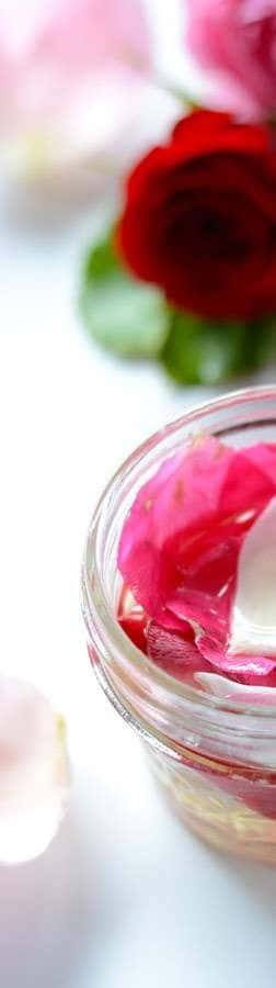 Rose Oil 桌布 壁紙 — 一個裝滿粉紅色玫瑰花瓣和液體的玻璃罐，放在明亮的白色表面上，旁邊是一朵鮮豔的紅玫瑰。