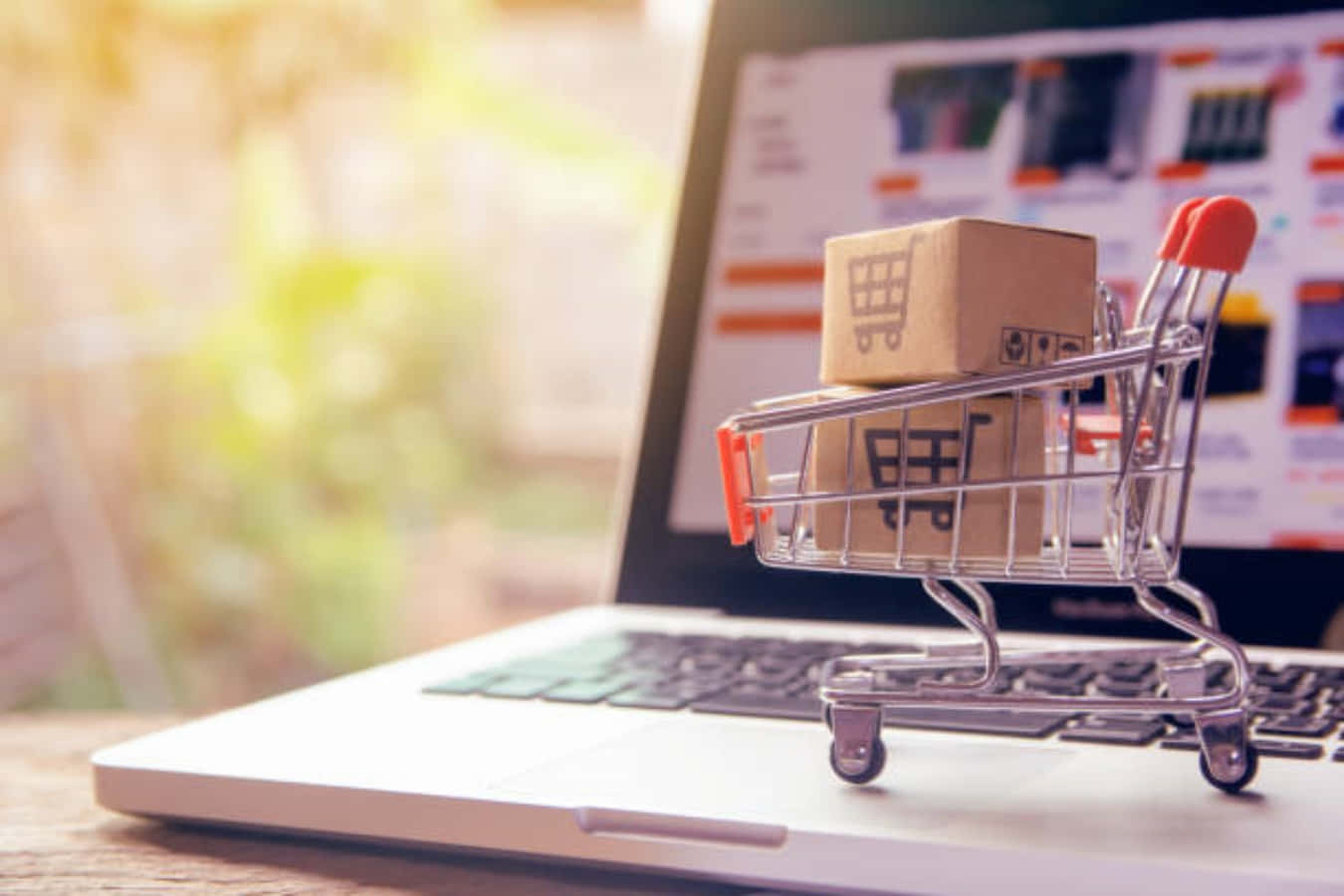 E Commerce 桌布 壁紙 — 一個裝滿紙箱的微型購物車放在一台顯示線上商店的筆記型電腦上，背景有模糊的綠色植物。
