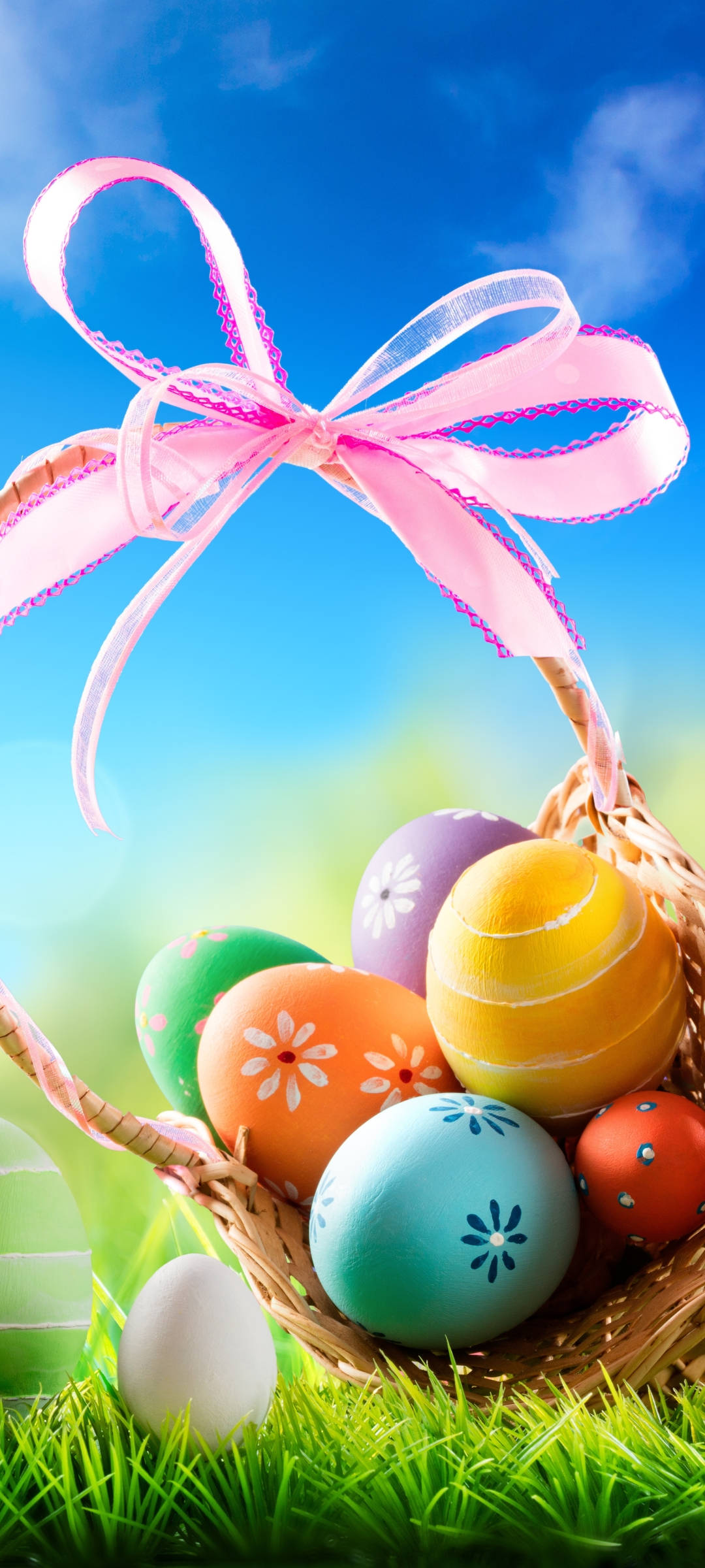 Easter Phone 桌布 壁紙 — 一個裝滿色彩鮮豔的復活節彩蛋和粉紅色絲帶的柳條籃子，放在茂密的綠草地上，頭頂是明亮的藍天。