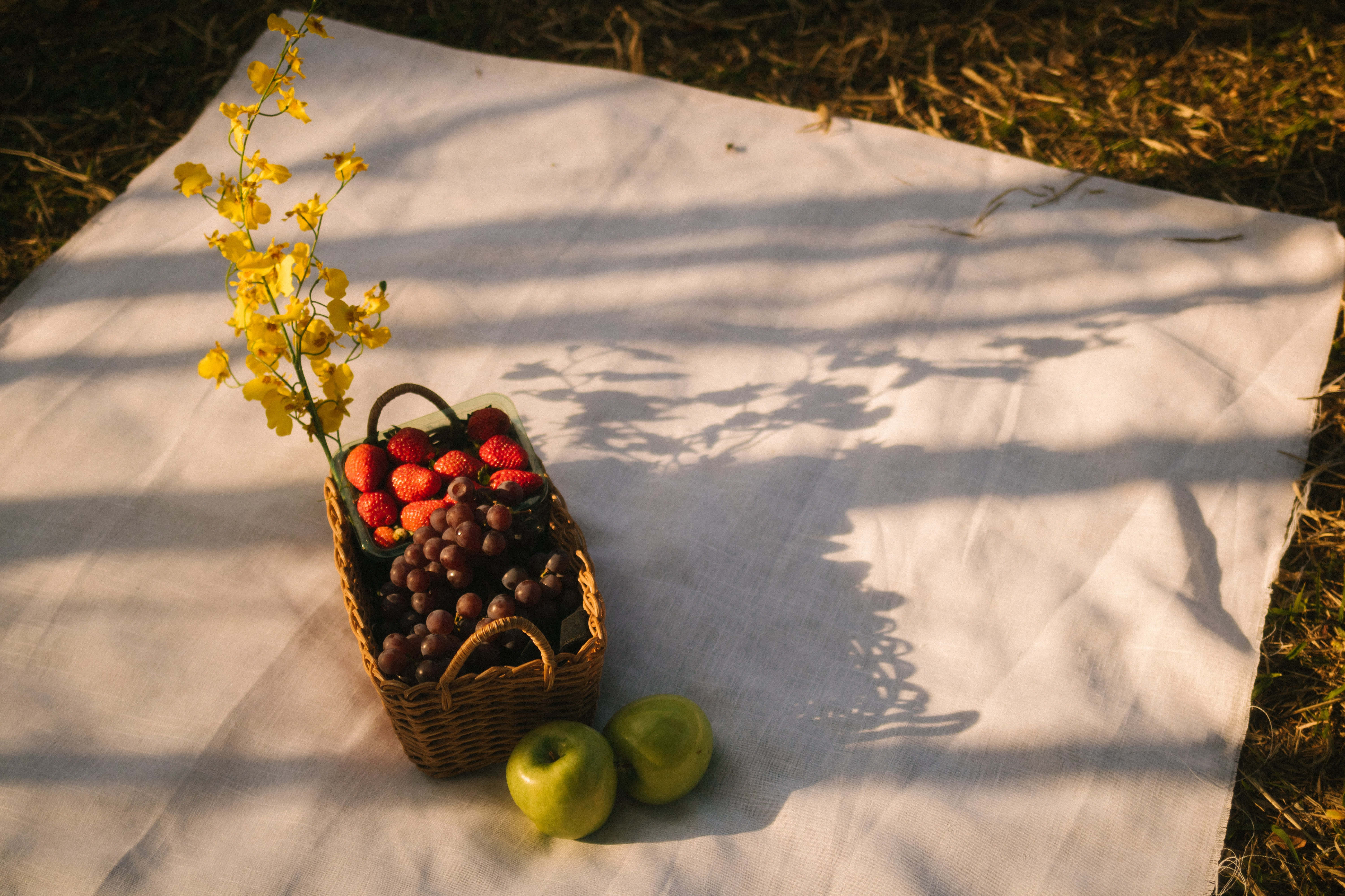 Summer Fruits 桌布 壁紙 — 一個裝滿草莓和葡萄的柳條籃子放在白色布上，旁邊有兩個青蘋果，斑駁的陽光灑落。