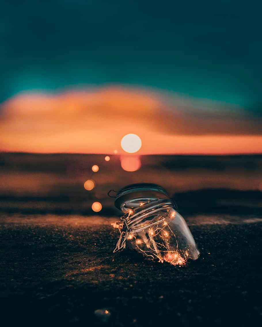 firefly 桌布 壁紙 — 一個裝滿閃爍仙女燈的玻璃瓶放在深色沙灘上，夕陽在火橙色和藍綠色的雲層中落下。