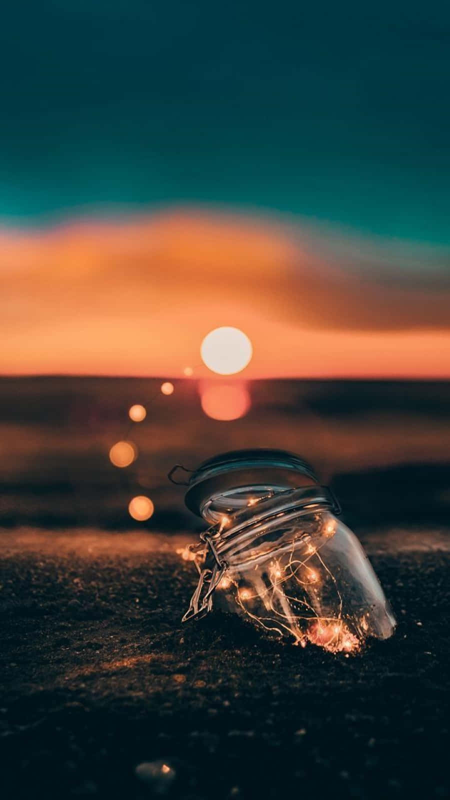 Sunset Photography 桌布 壁紙 — 一個裝滿閃爍仙女燈的玻璃瓶放在深色沙灘上，夕陽在火橙色和青綠色的天空中落下。