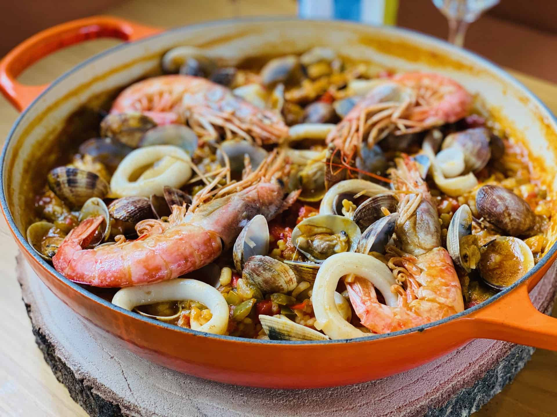 prawn 桌布 壁紙 — 一個裝滿鮮美蝦、蛤蜊和魷魚的鮮橙色西班牙海鮮飯鍋，放置在質樸的木板上。