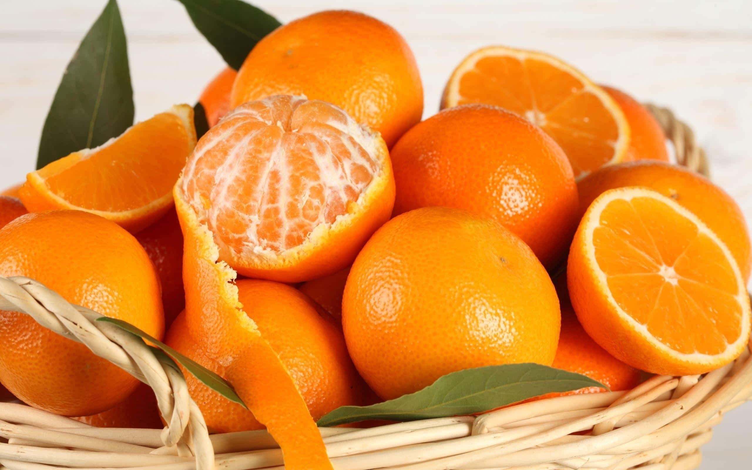 Orange Fruit 桌布 壁紙 — 一個裝滿鮮豔橙子的籃子，有些完整，有些剝了皮，露出多汁的果瓣和新鮮的綠葉。