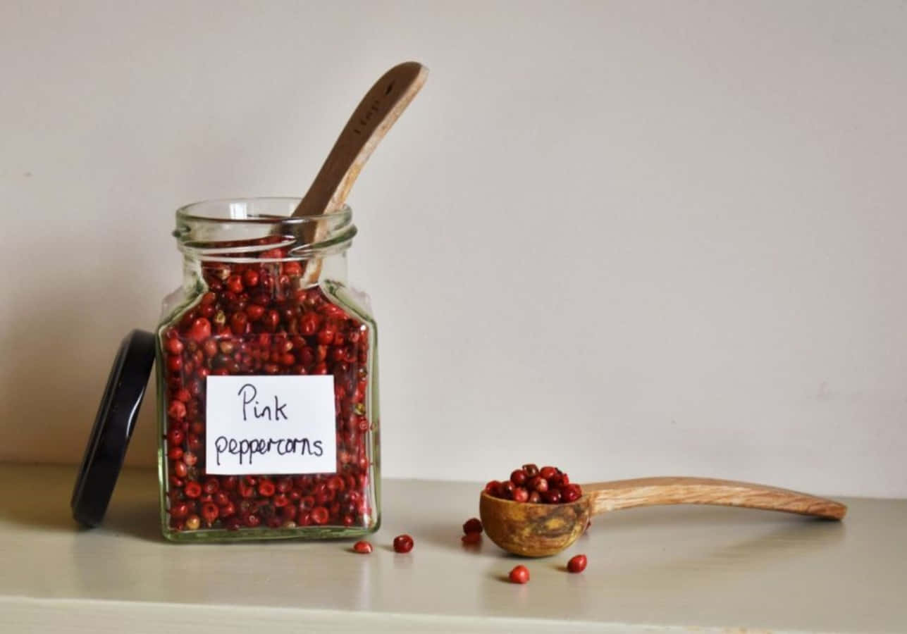 Pink Peppercorn 桌布 壁紙 — 一個裝滿鮮豔粉紅胡椒粒的玻璃罐，旁邊放著一把木勺，上面還有更多胡椒粒，放在淺色檯面上。
