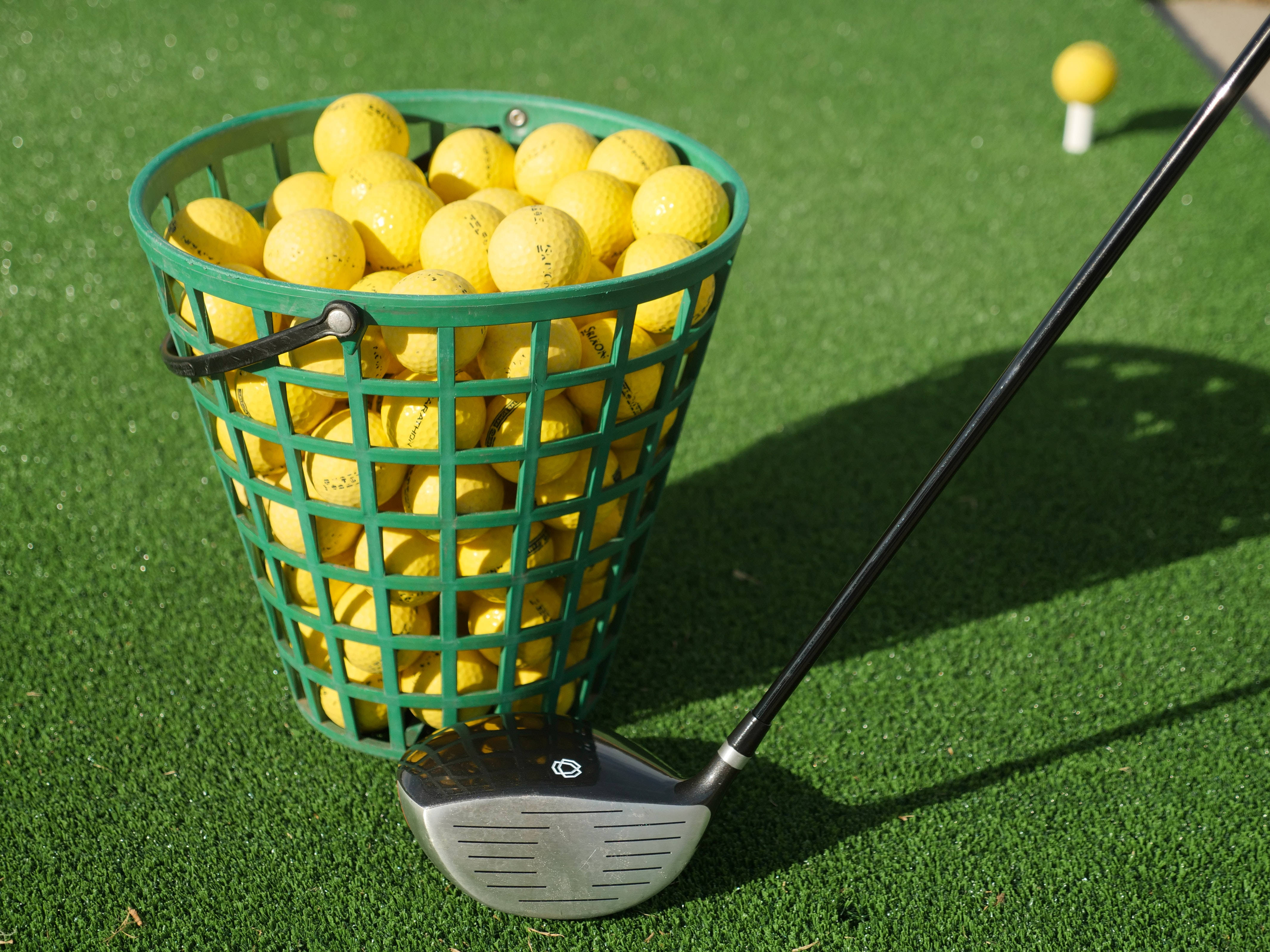 4K Golf 桌布 壁紙 — 一個裝滿鮮黃色高爾夫球的綠色籃子，在晴朗的陽光下，放在翠綠的草地上，旁邊有一支高爾夫球桿。