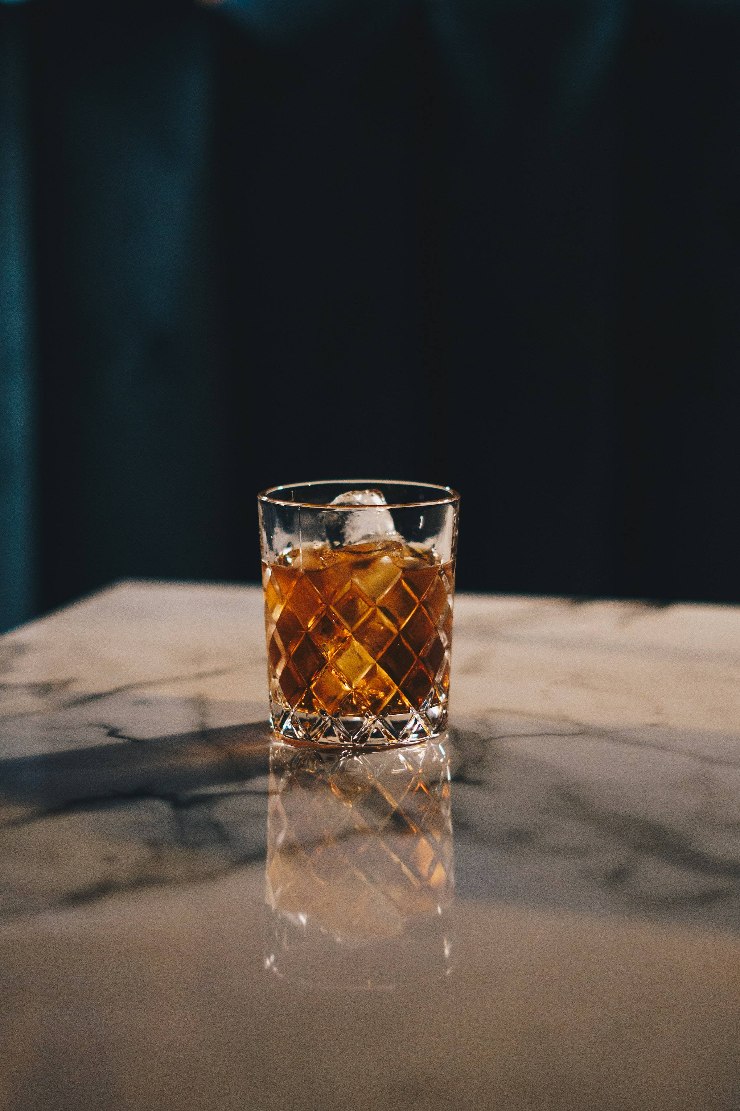 alcohol 桌布 壁紙 — 一個裝著琥珀色威士忌和冰塊的水晶杯放在拋光的大理石表面上，反射出溫暖的光線，背景是深藍綠色。