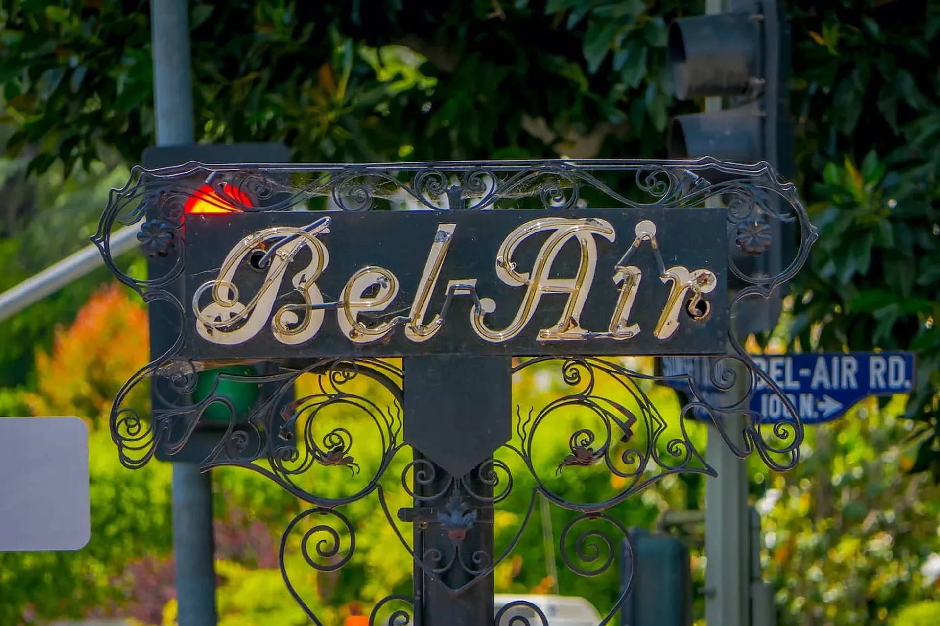 Boulevard 桌布 壁紙 — 一個裝飾性的黑色鍛鐵標誌，帶有金色 'Bel-Air' 字樣，矗立在街角，周圍環繞著茂密的綠色樹葉。