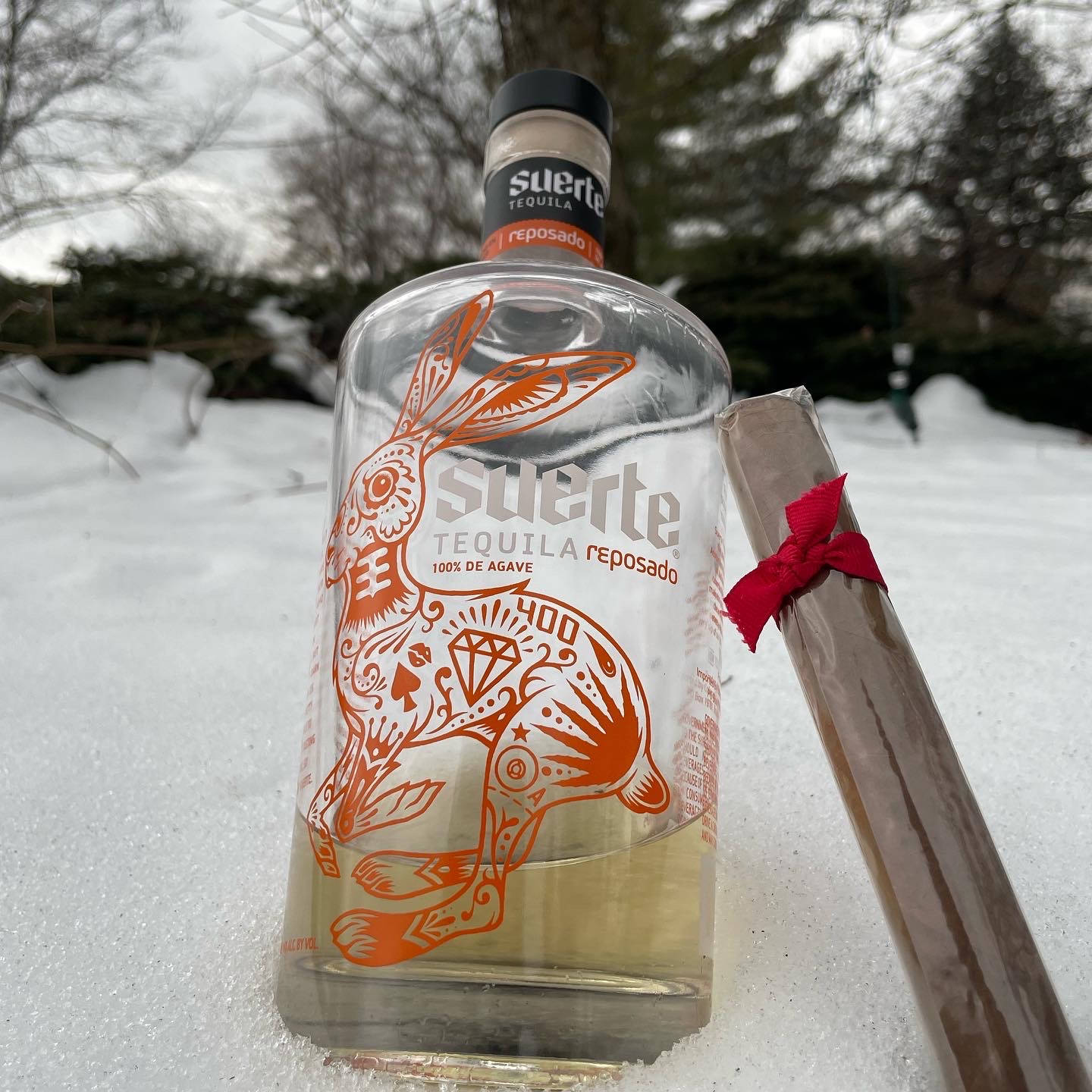 bottle 桌布 壁紙 — 一個裝飾性的龍舌蘭酒瓶，帶有橙色兔子設計，放在新鮮的雪中，旁邊有一支包裹著的雪茄。