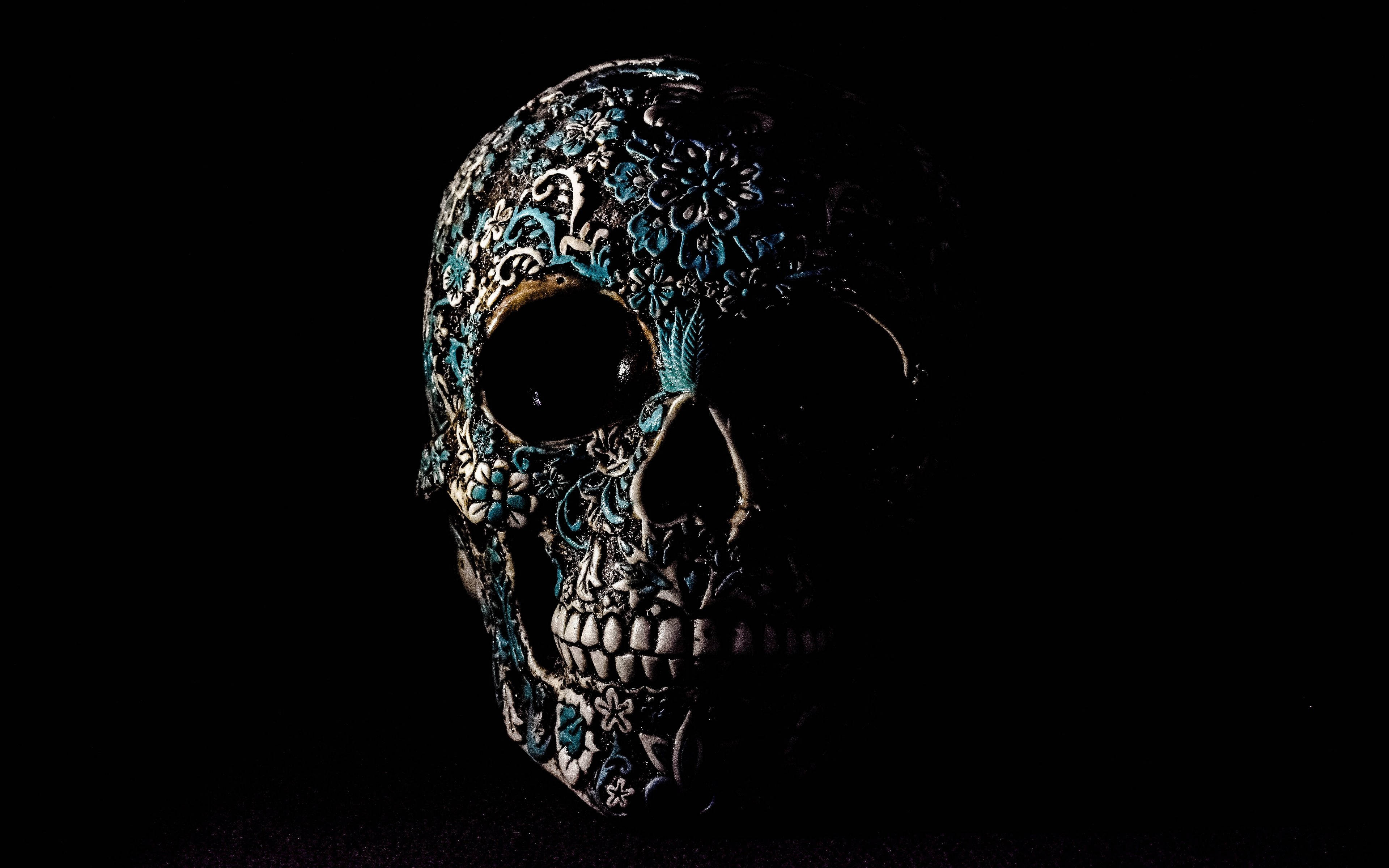 Black Skull 桌布 壁紙 — 一個裝飾精美的頭骨，帶有藍色和銀色花卉圖案，在漆黑的背景下發光。