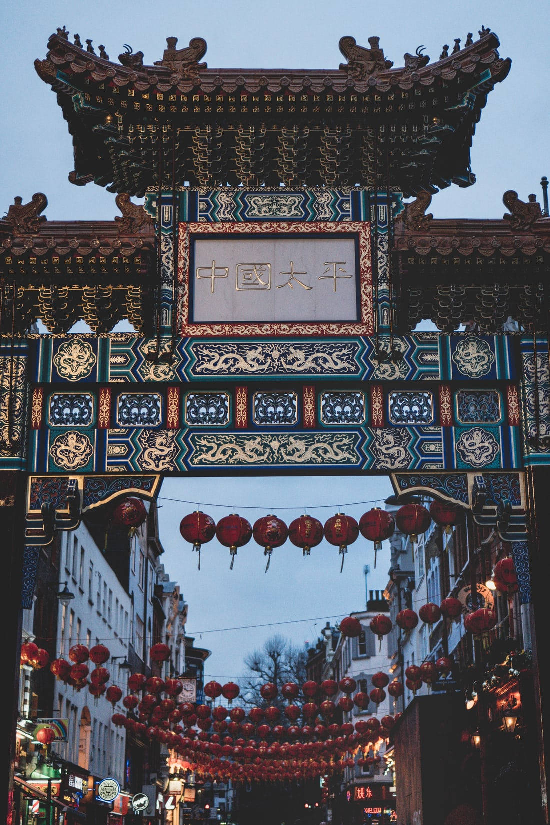 唐人街 桌布 壁紙 — 一個裝飾華麗的傳統中式牌坊，掛著紅燈籠，懸在黃昏時分的狹窄街道上。