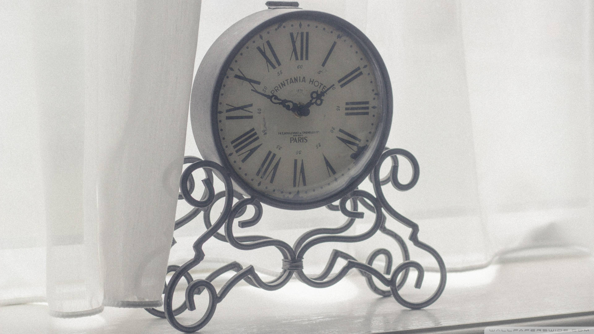 Time 桌布 壁紙 — 一個裝飾華麗的復古時鐘，帶有羅馬數字，放置在精緻的金屬架上，旁邊是輕柔的窗簾，在柔和的日光中。