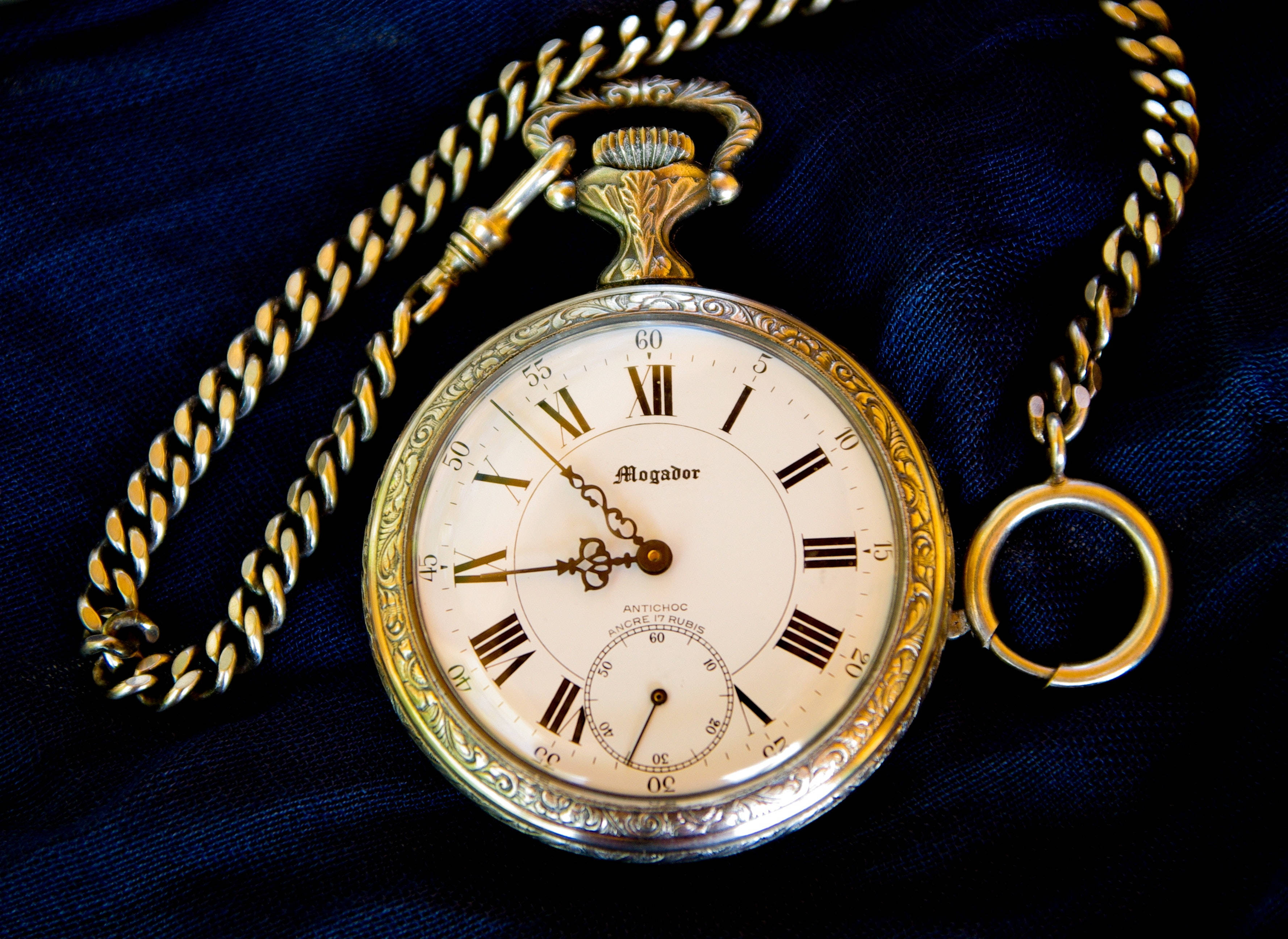 Gold Jewellery 桌布 壁紙 — 一個裝飾華麗的金色懷錶，帶有羅馬數字，放在濃郁的藍色天鵝絨布上，鏈條盤繞在旁邊。