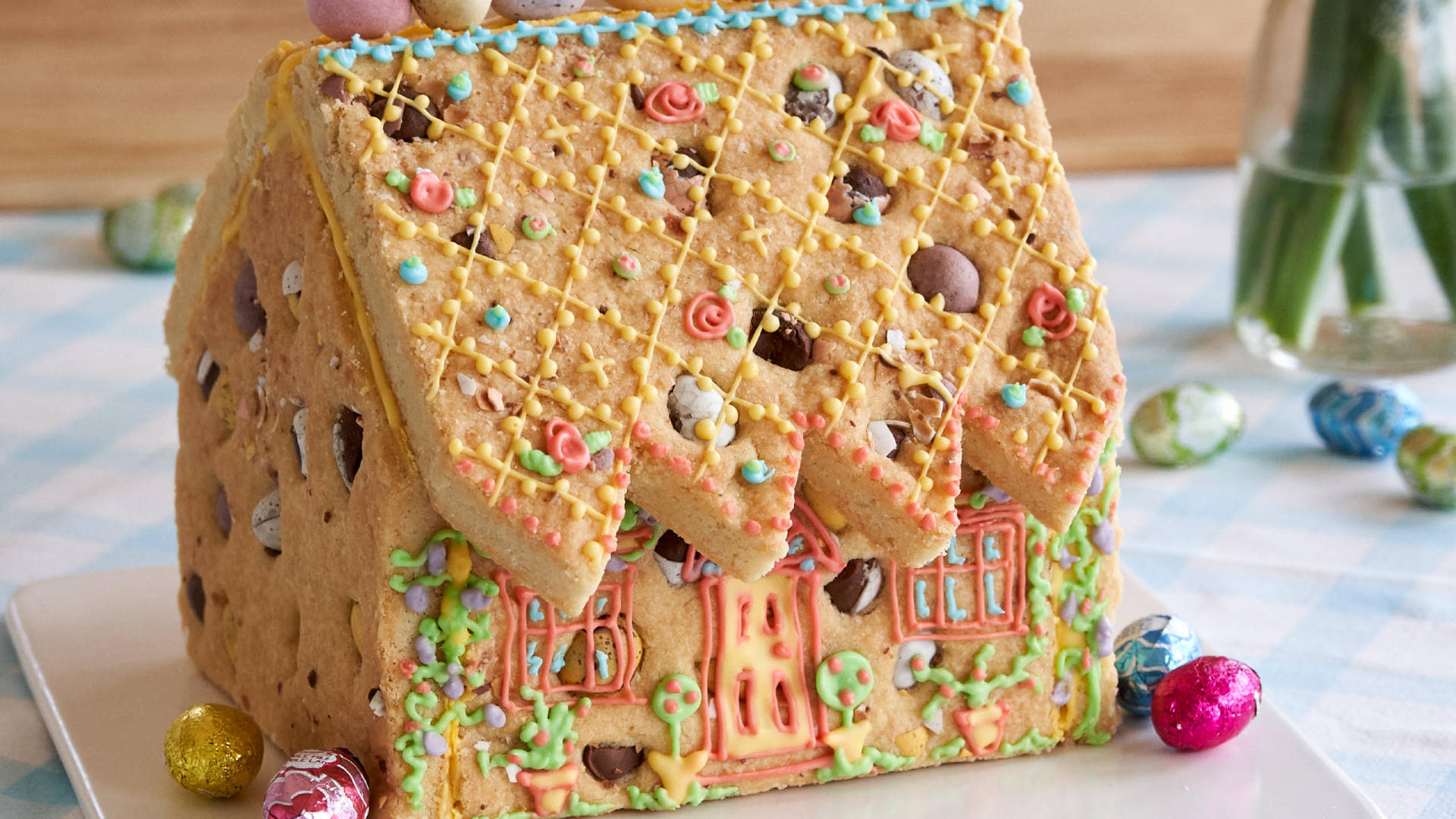 Gingerbread House 桌布 壁紙 — 一個裝飾著彩色糖霜花、巧克力豆和糖果蛋的夢幻薑餅屋，放在格子桌布上。