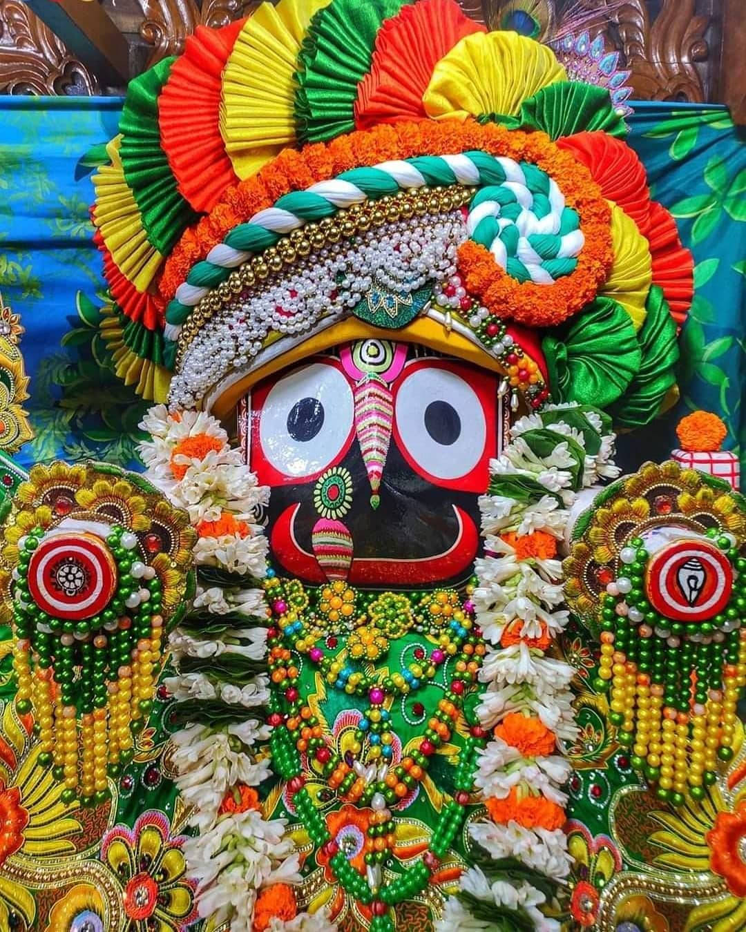 Jagannath 桌布 壁紙 — 一個裝飾鮮豔的印度教神像，飾有彩色紙扇、珠子、花朵和精緻的珠寶，背景為圖案背景。