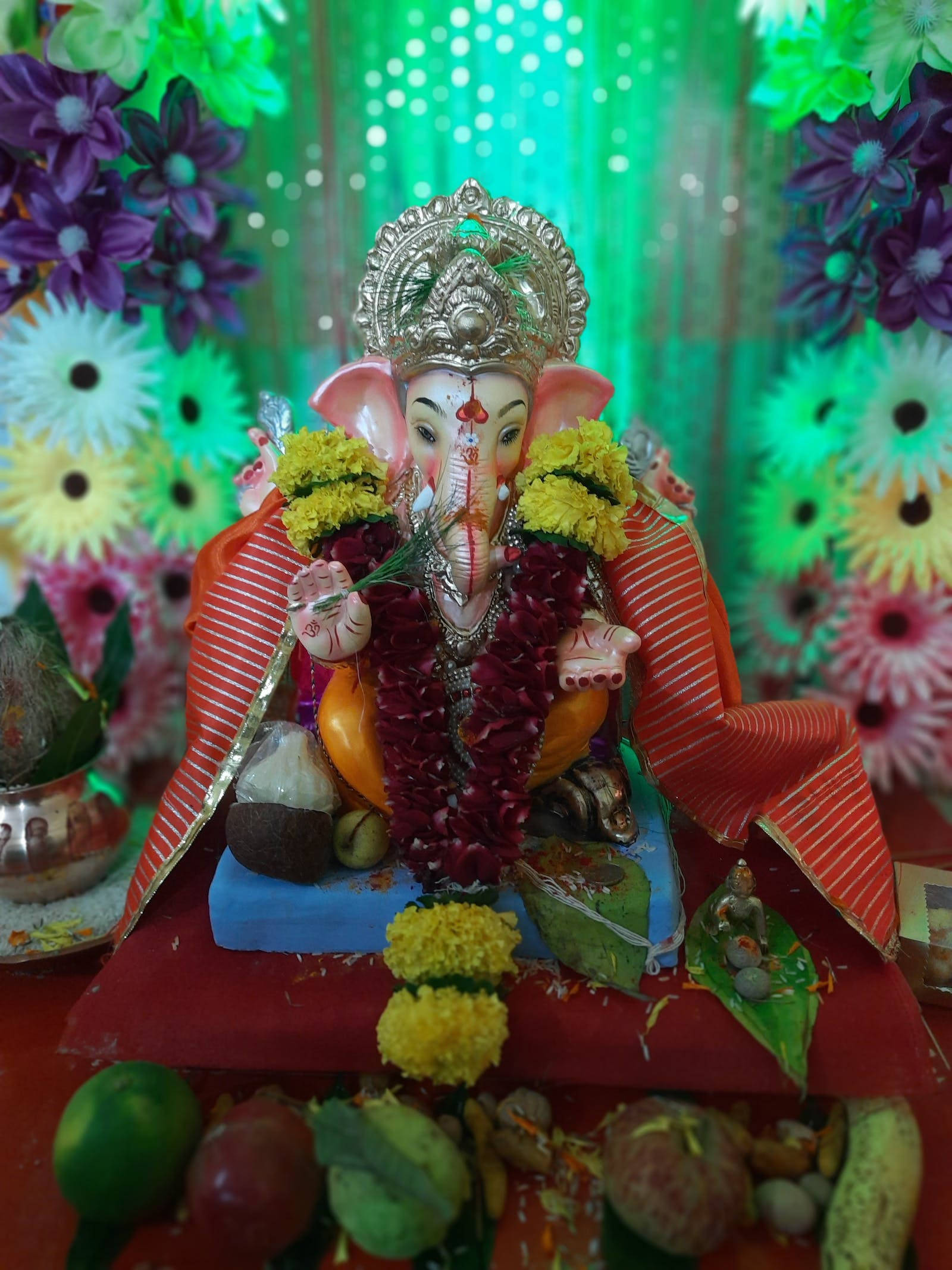 Ganesh 桌布 壁紙 — 一個裝飾鮮豔的象神像，飾有萬壽菊和紅色花環，坐在一塊紅布上，周圍環繞著色彩繽紛的花朵和供品。