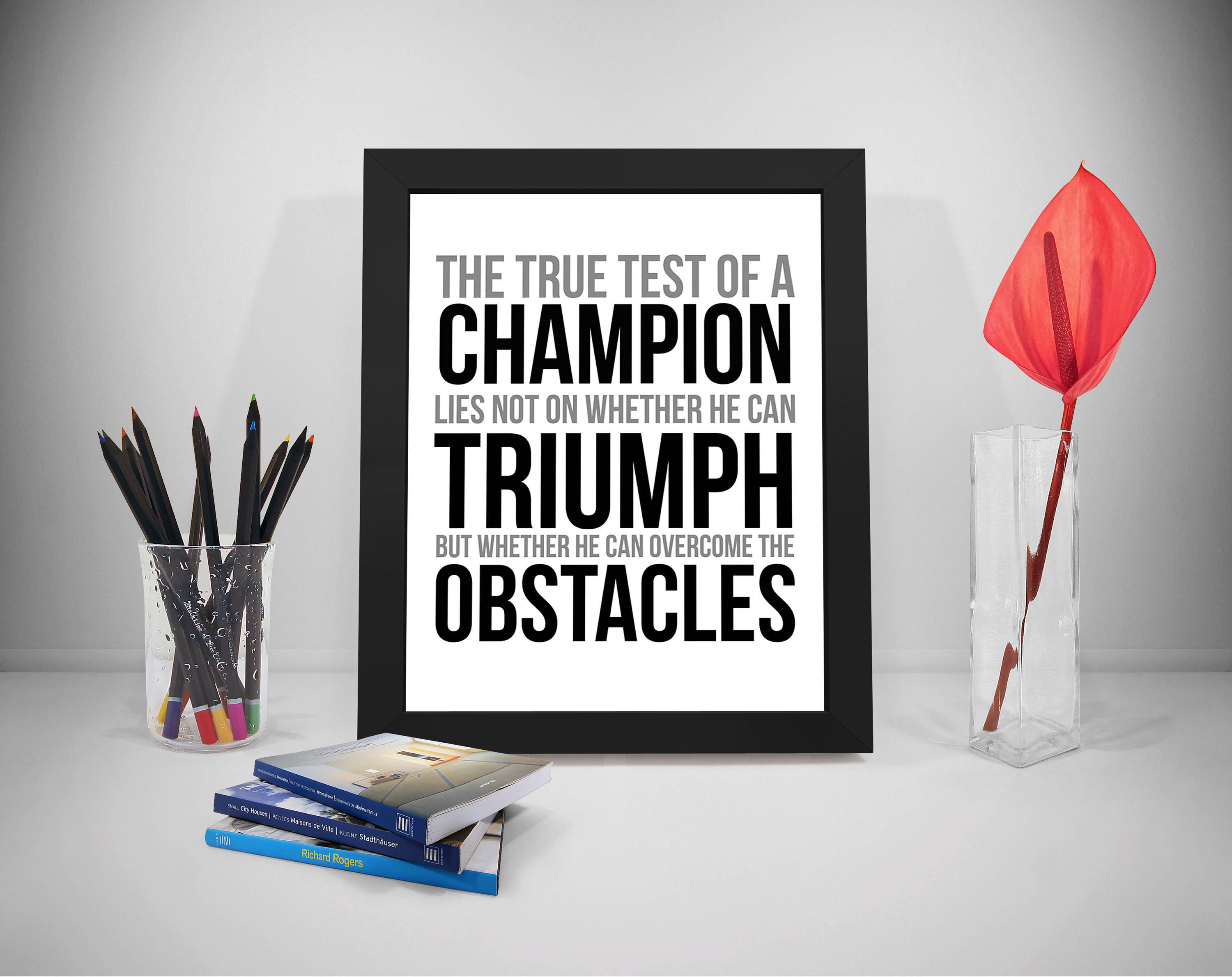Champion 桌布 壁紙 — 一個裱框的勵志名言，關於冠軍克服障礙，放在乾淨的白色書桌上，旁邊有書本、彩色鉛筆和一個玻璃花瓶中的紅花。