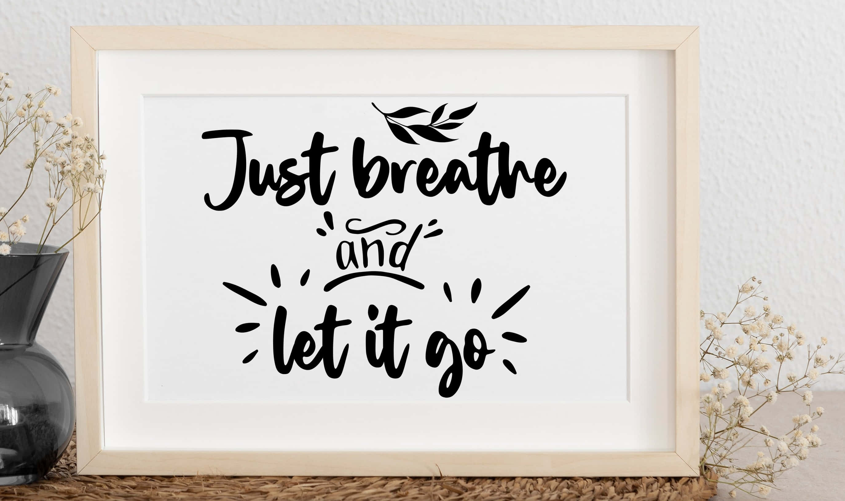 語錄 桌布 壁紙 — 一個裱框的勵志語錄，上面用黑色手寫體寫著「Just breathe and let it go」，並有葉子裝飾，兩側是放在編織表面花瓶中的乾燥滿天星。