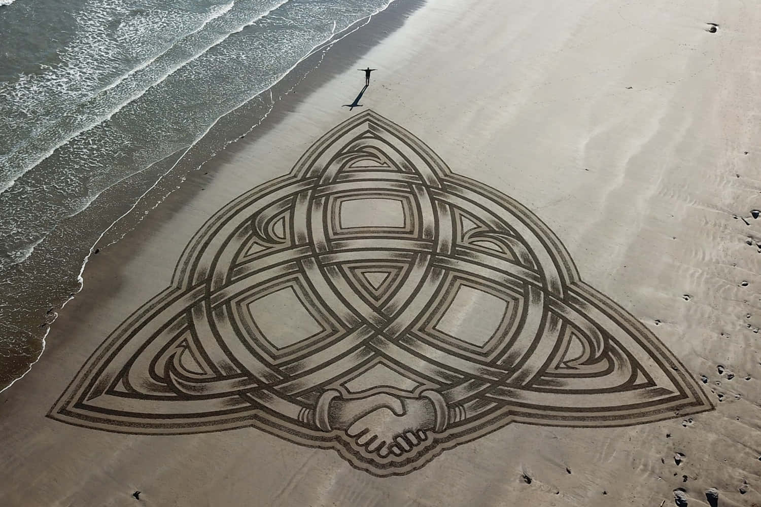 Sand Art 桌布 壁紙 — 一個複雜的凱爾特結沙藝設計，帶有握手圖案，繪製在濕漉漉的海灘上，海浪翻滾而來。