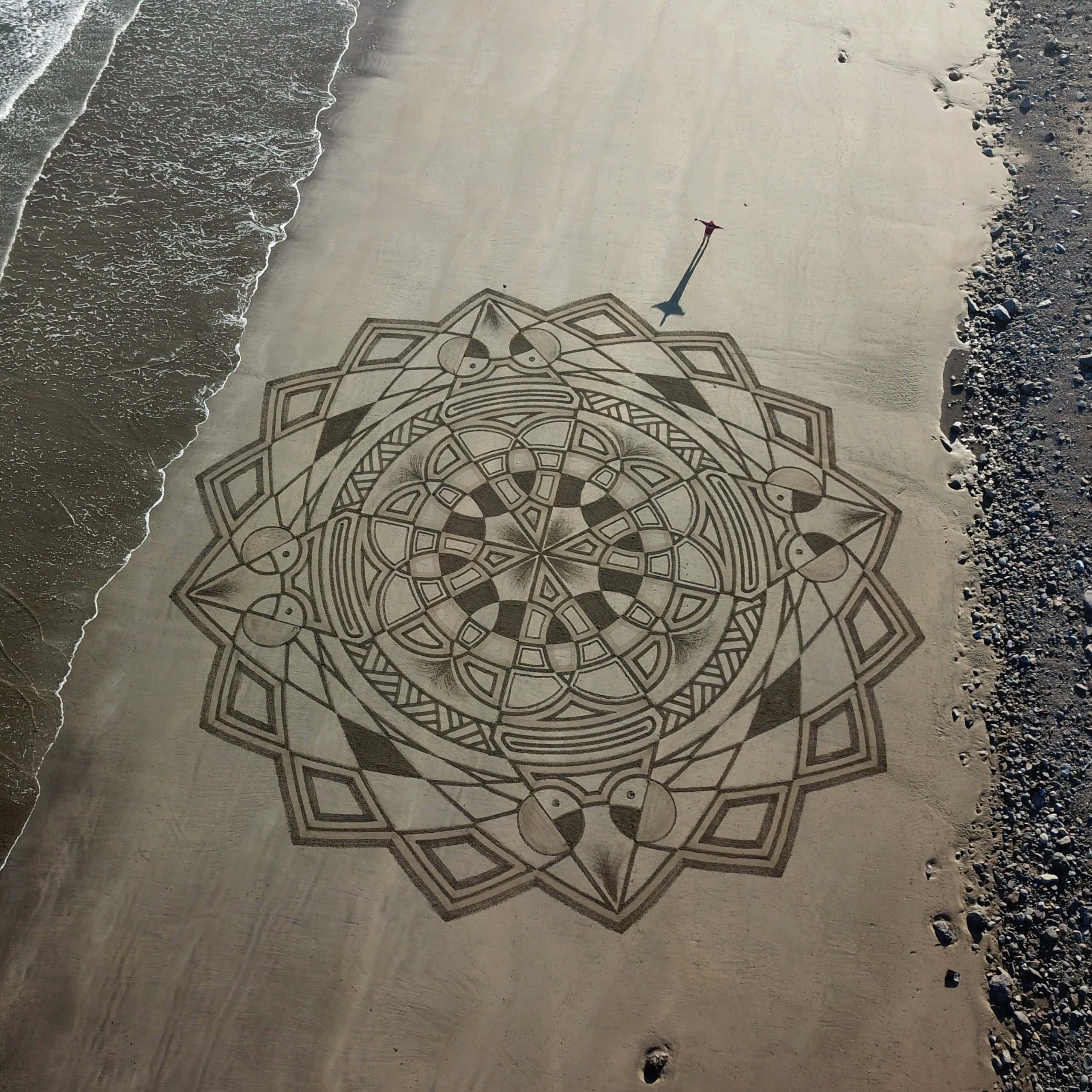 Sand Art 桌布 壁紙 — 一個複雜的幾何曼陀羅刻在海岸線的濕沙上，溫柔的海浪拍打著它的邊緣。