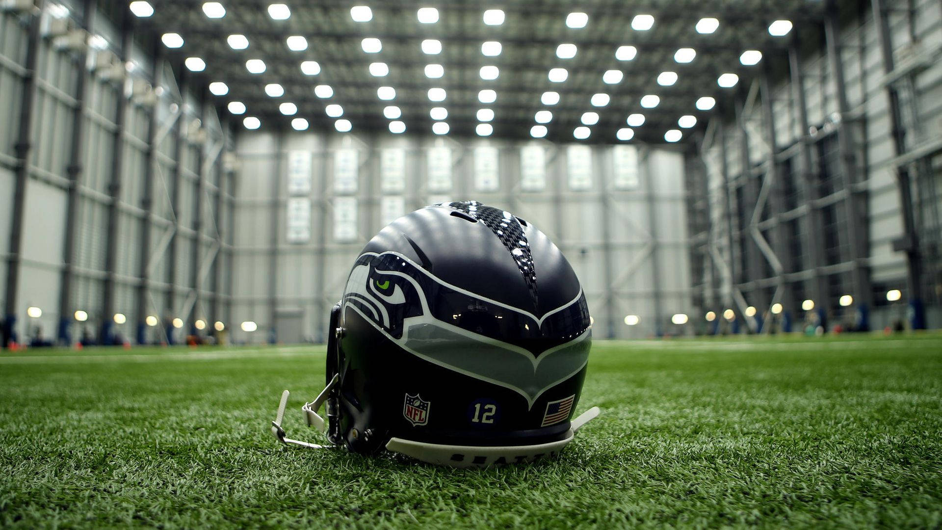 Seattle Seahawks 桌布 壁紙 — 一個西雅圖海鷹隊的美式足球頭盔放在廣闊、光線充足的室內體育場的鮮綠色草地上。